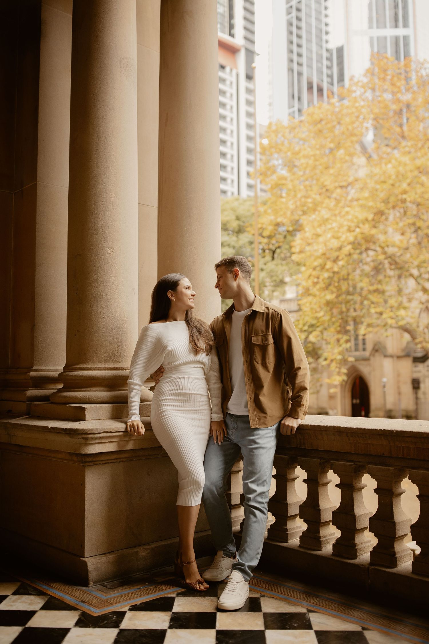 Stylish couple sharing intimate moment on historic balcony with autumn trees in background.