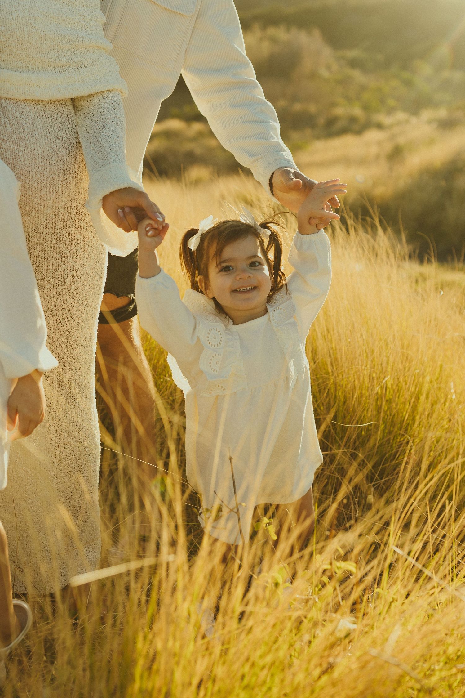 Family members dressed in white hold hands while standing in golden sunlit grass during sunset.