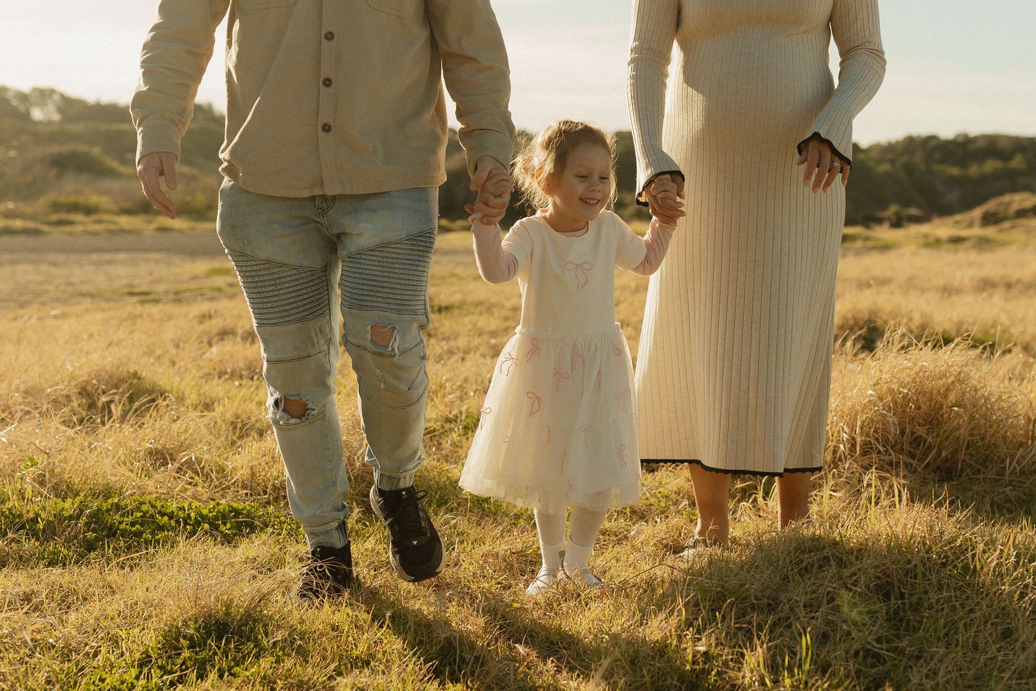 Family walks together through golden grass at sunset with child holding hands between parents.