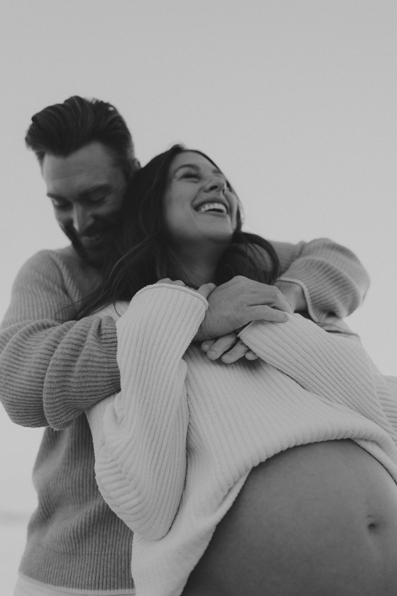 Black and white photo of a couple embracing on a beach, man hugging pregnant woman from behind as she laughs joyfully.