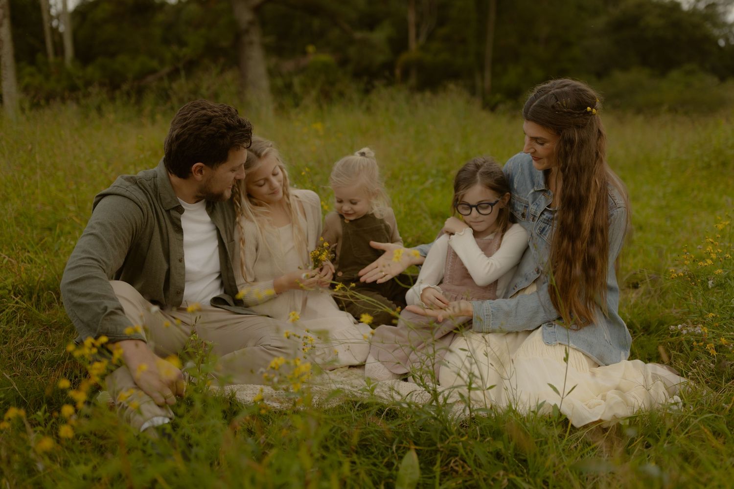 Family of five enjoying a peaceful picnic in a lush green meadow with wildflowers on a warm day.