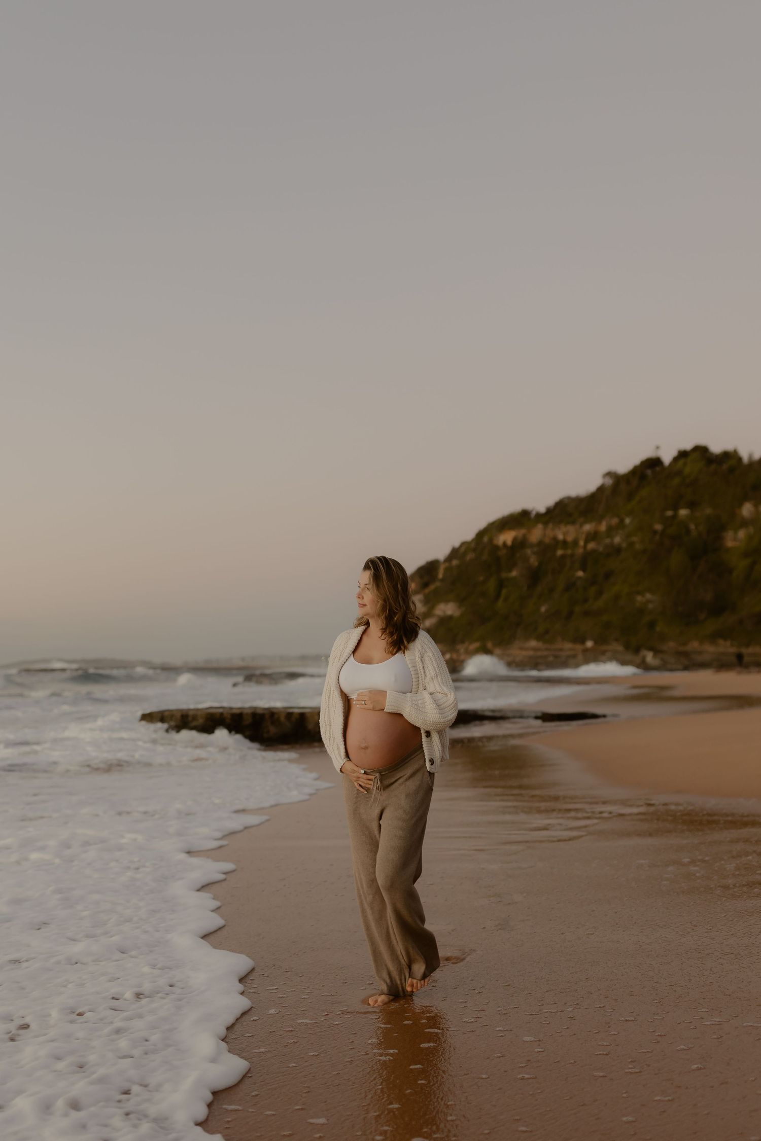 Pregnant woman in white cardigan and tan pants walking along sandy beach at sunset near rocky headland.