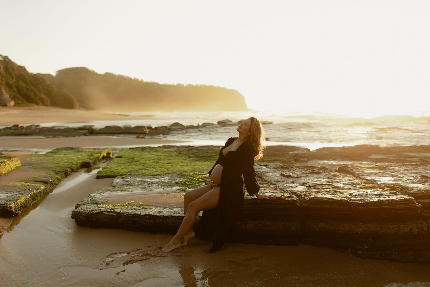 Woman in black dress sitting on mossy rocks at sunset by the ocean with golden light and waves in the background.