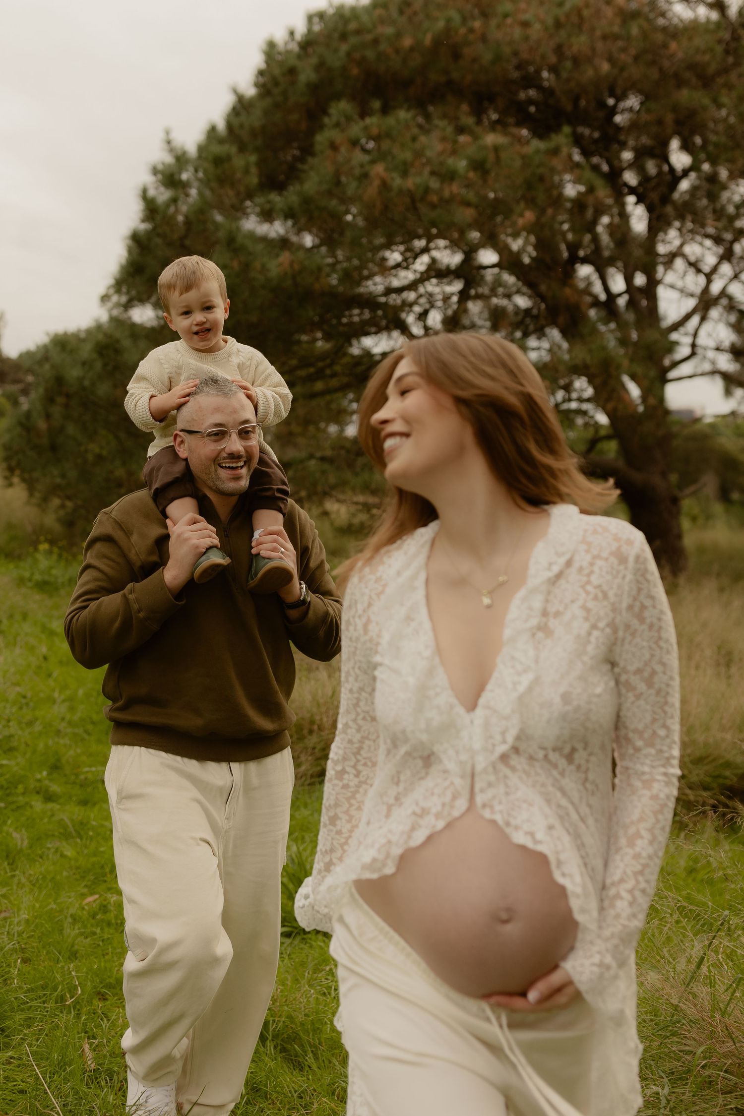 Pregnant woman in white lace dress smiles as man carries toddler on shoulders in green field.