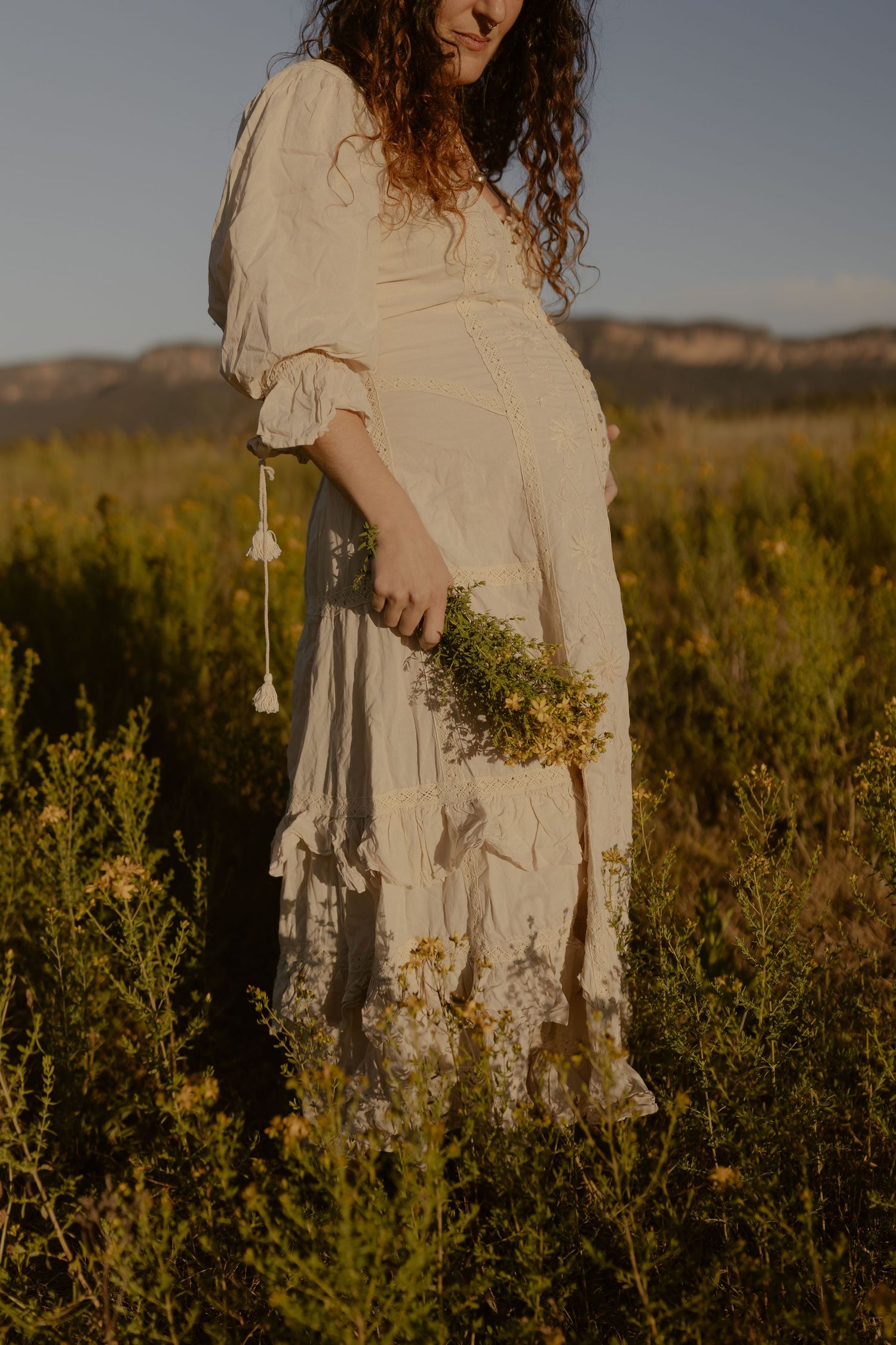 A flowing white dress catches golden light as wildflowers are held in a meadow at sunset.