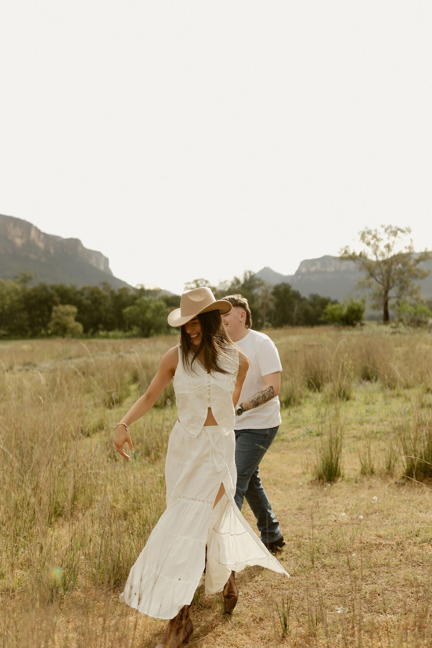 A couple dances playfully in a field with mountains in the background at golden hour.