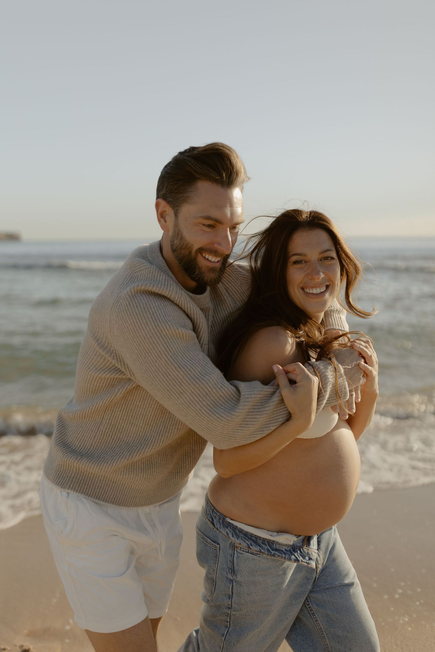 Playful couple embraces and laughs together on a sunny beach with ocean waves in the background.