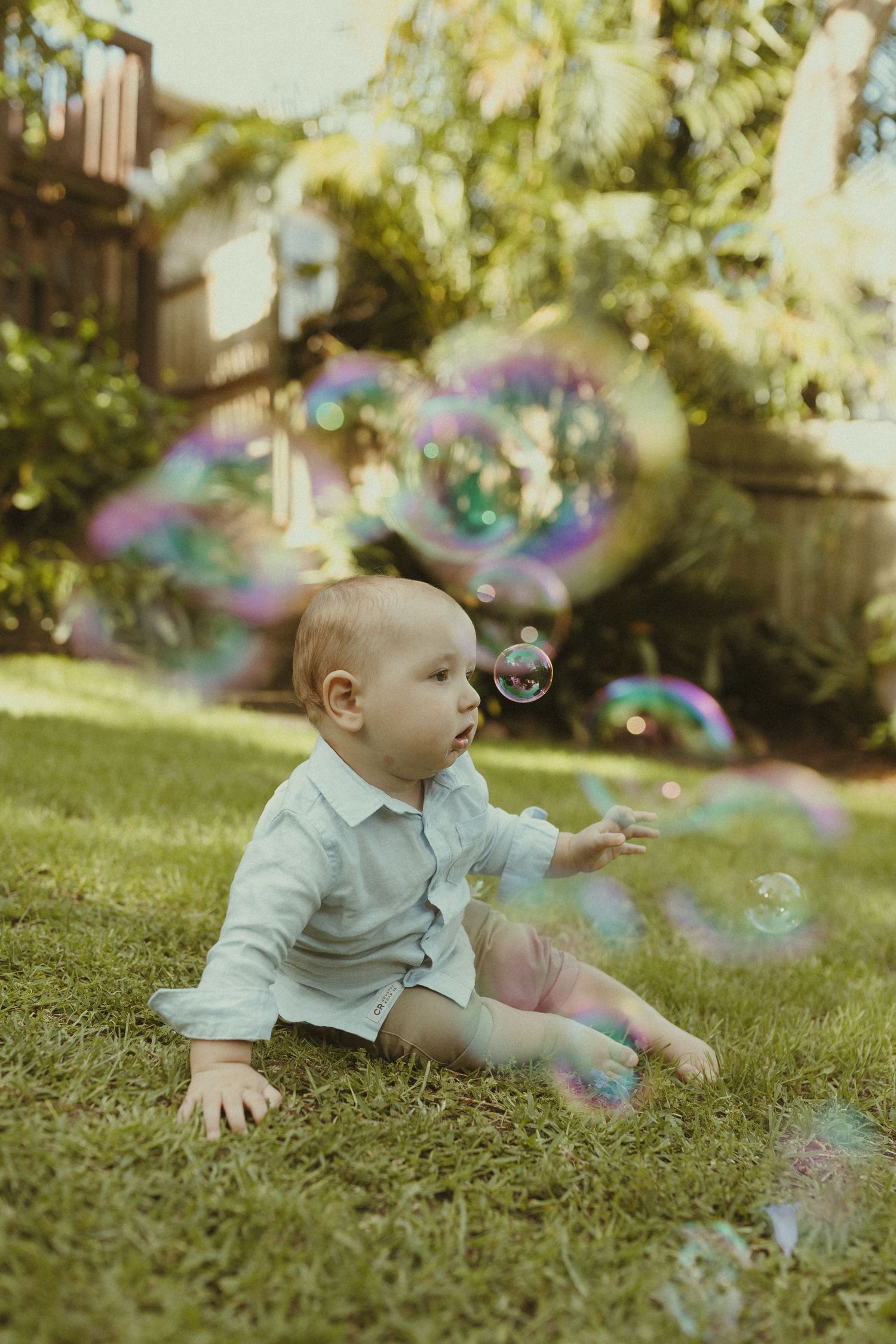 A baby in a light colored shirt crawls on grass while reaching for floating soap bubbles in a sunlit garden.