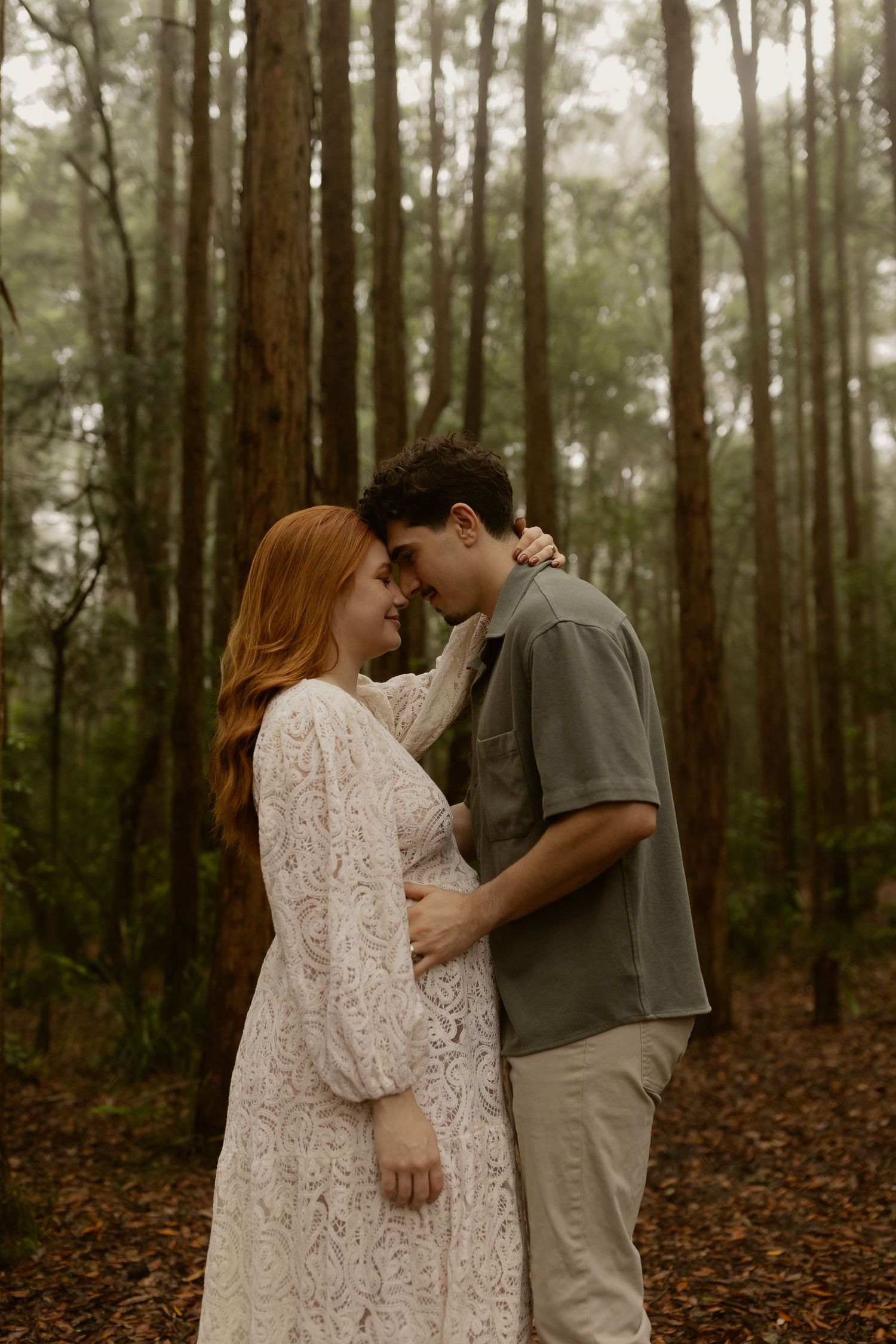 A couple embraces tenderly during a maternity session among tall trees in a forest setting.