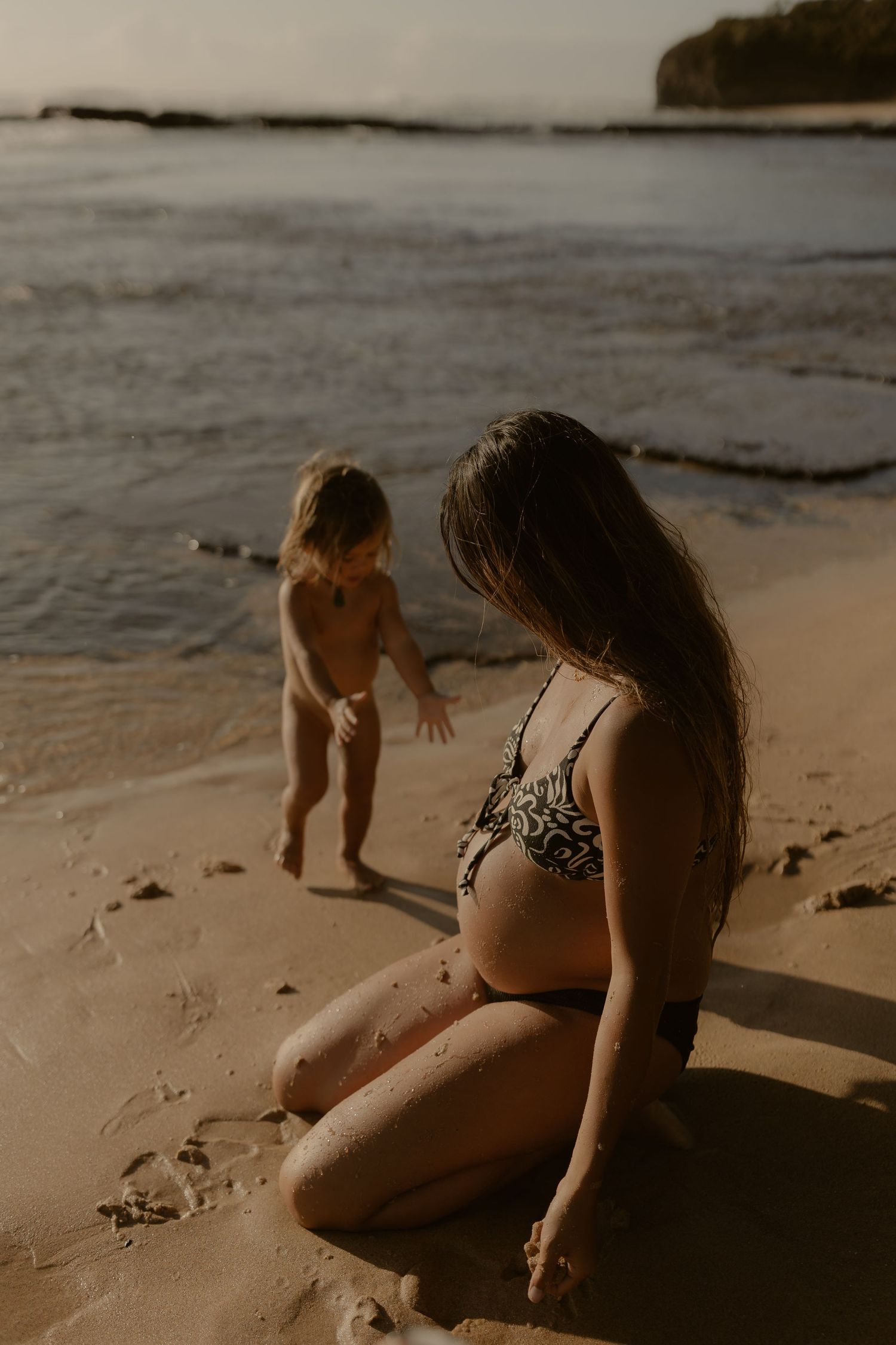 A mother and toddler play together on a sandy beach at sunset, creating a warm and intimate scene.