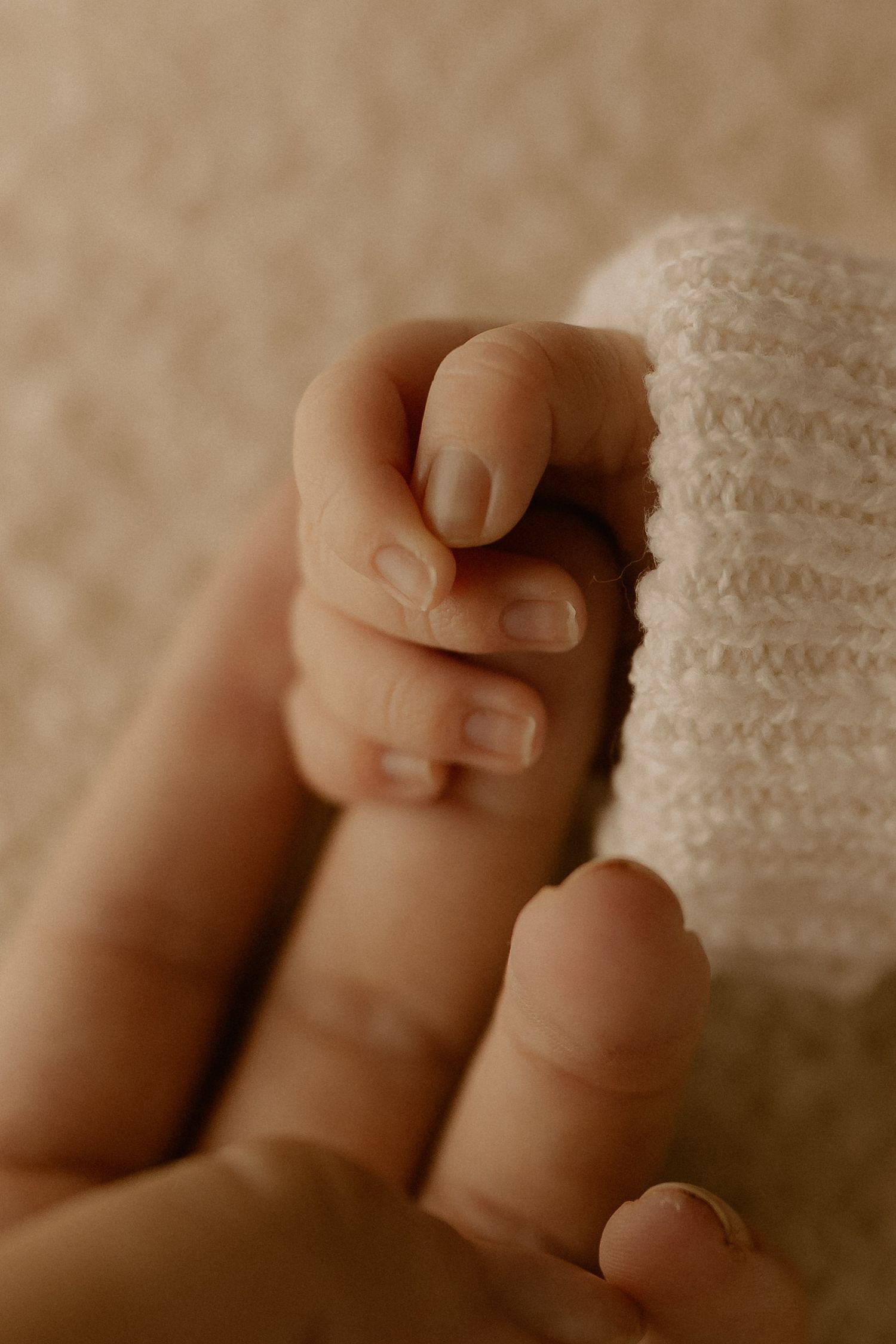 Close-up photo of tiny baby fingers grasping gently against soft white knitted fabric in warm sepia tones.