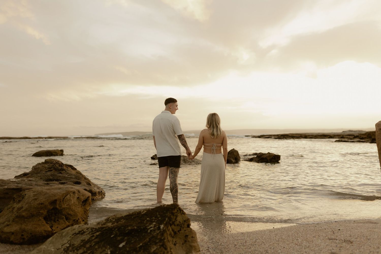 Couple walking hand in hand along a rocky beach at golden hour.