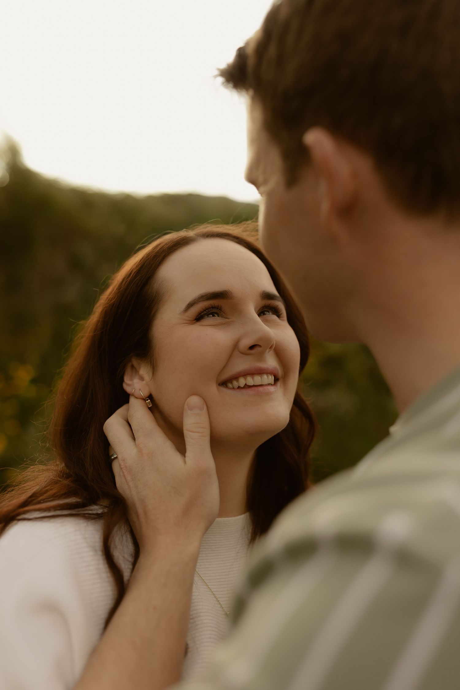 A smiling woman with auburn hair gazes up at a man gently cupping her face outdoors at golden hour.