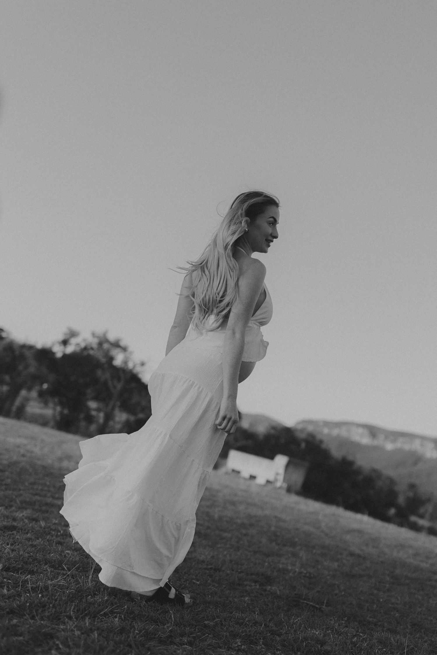 Black and white photo of woman in flowing white dress turning gracefully in open field.