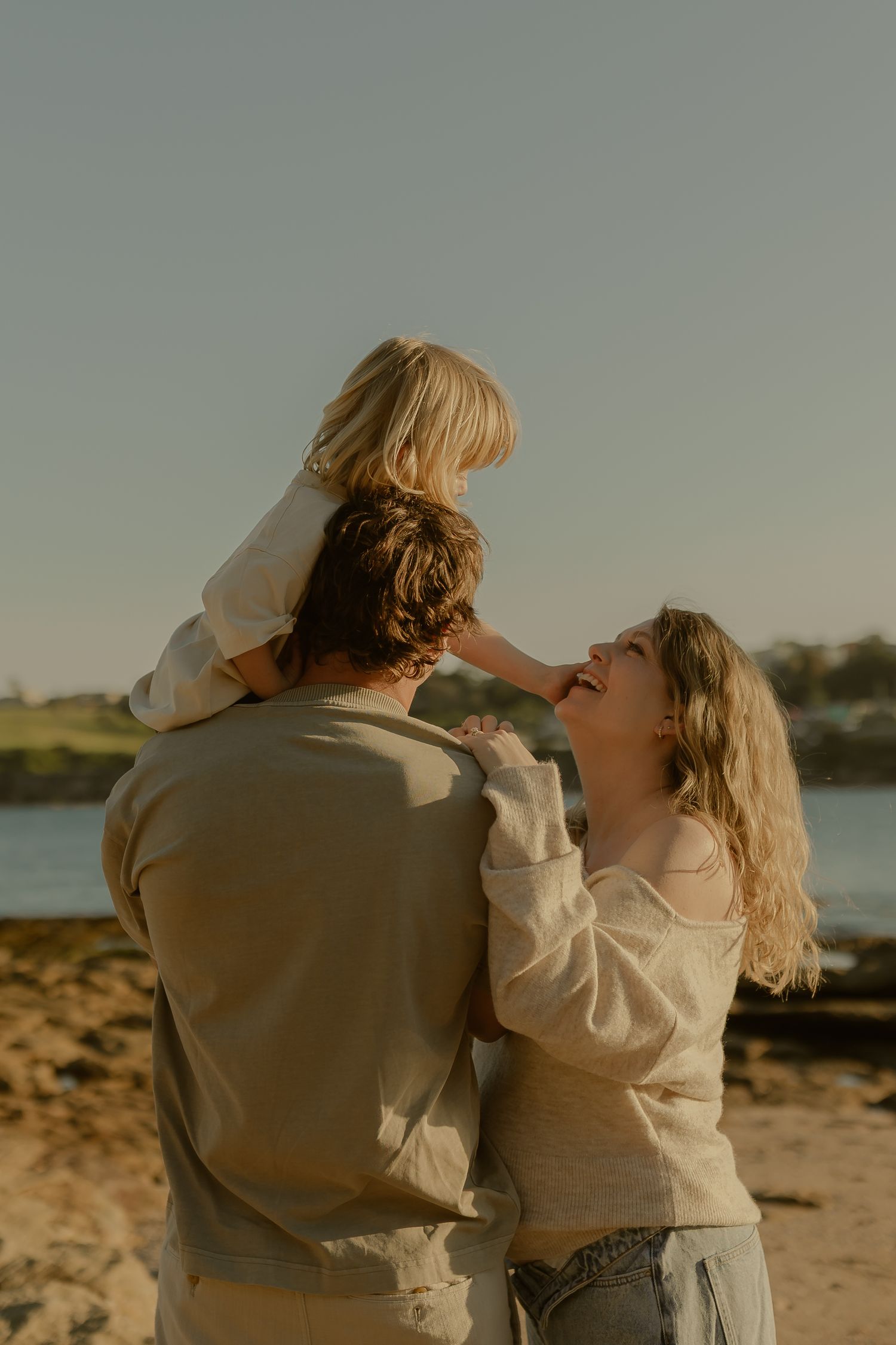 A joyful family at the beach, with a child sitting on dad's shoulders while mom laughs and reaches up lovingly.