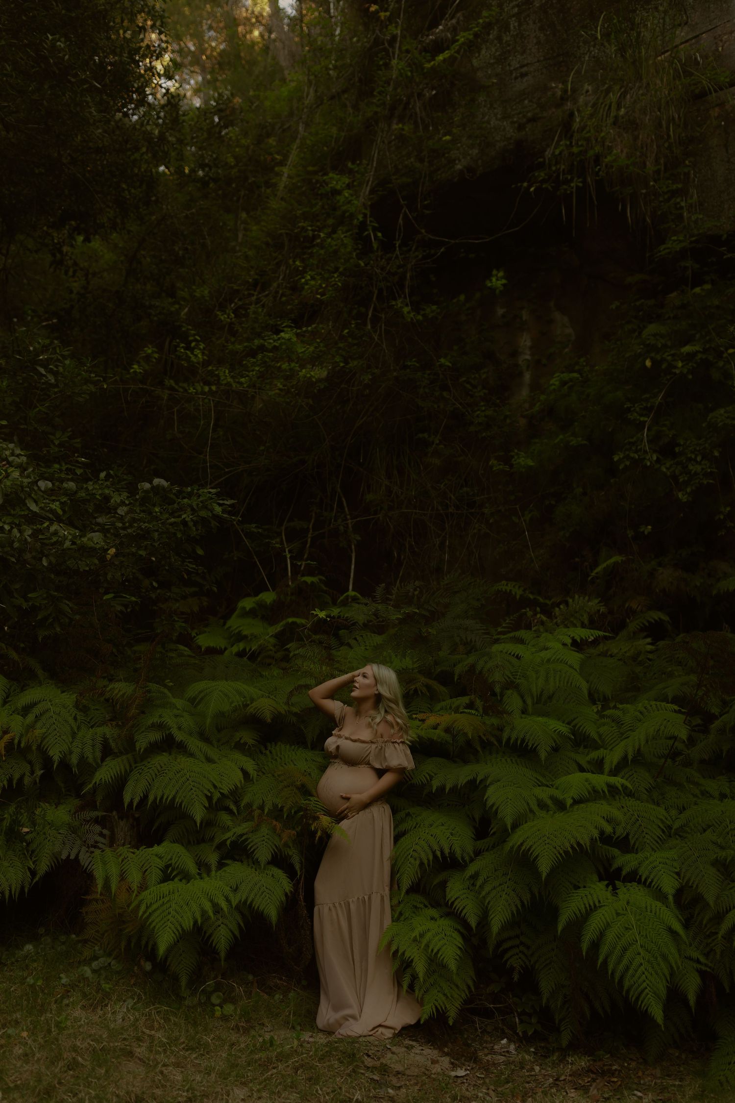 Pregnant woman in beige gown standing among lush ferns in a dark forest, cradling her baby bump.