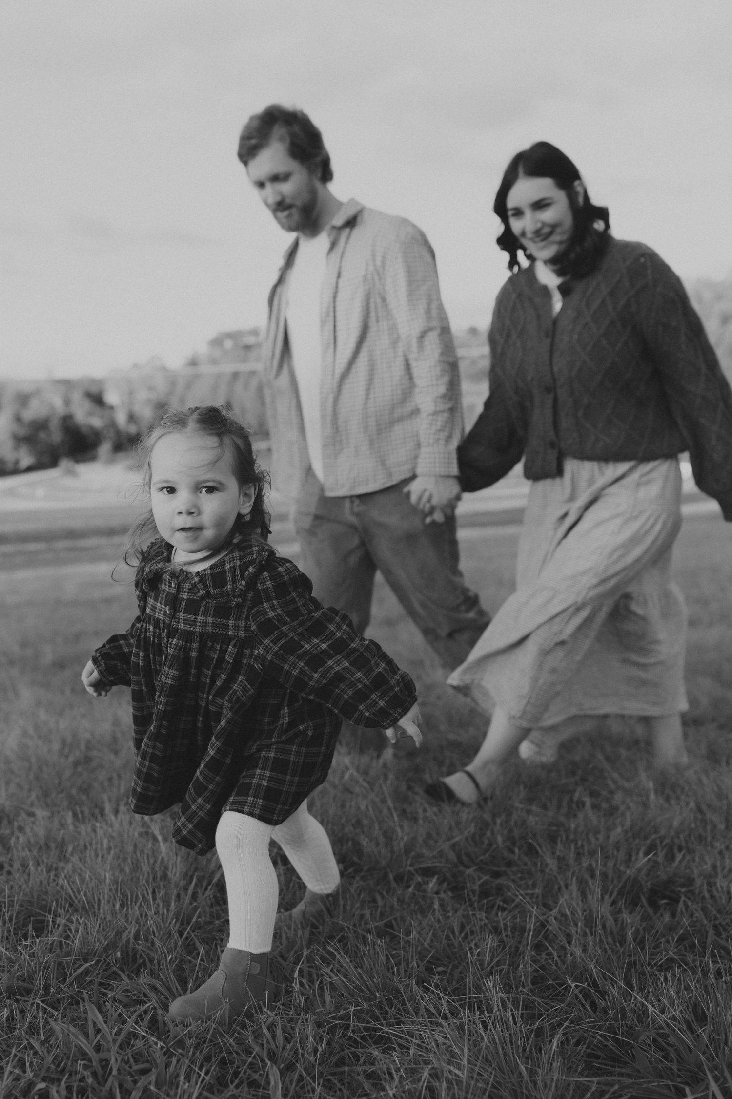 Black and white photo of a young family with a toddler girl running in a grassy field outdoors.