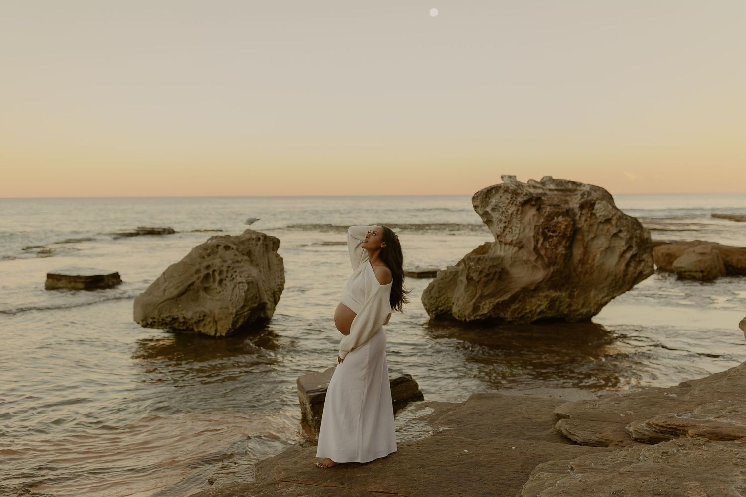 Pregnant woman in white dress standing by ocean rocks at sunset on a peaceful beach.