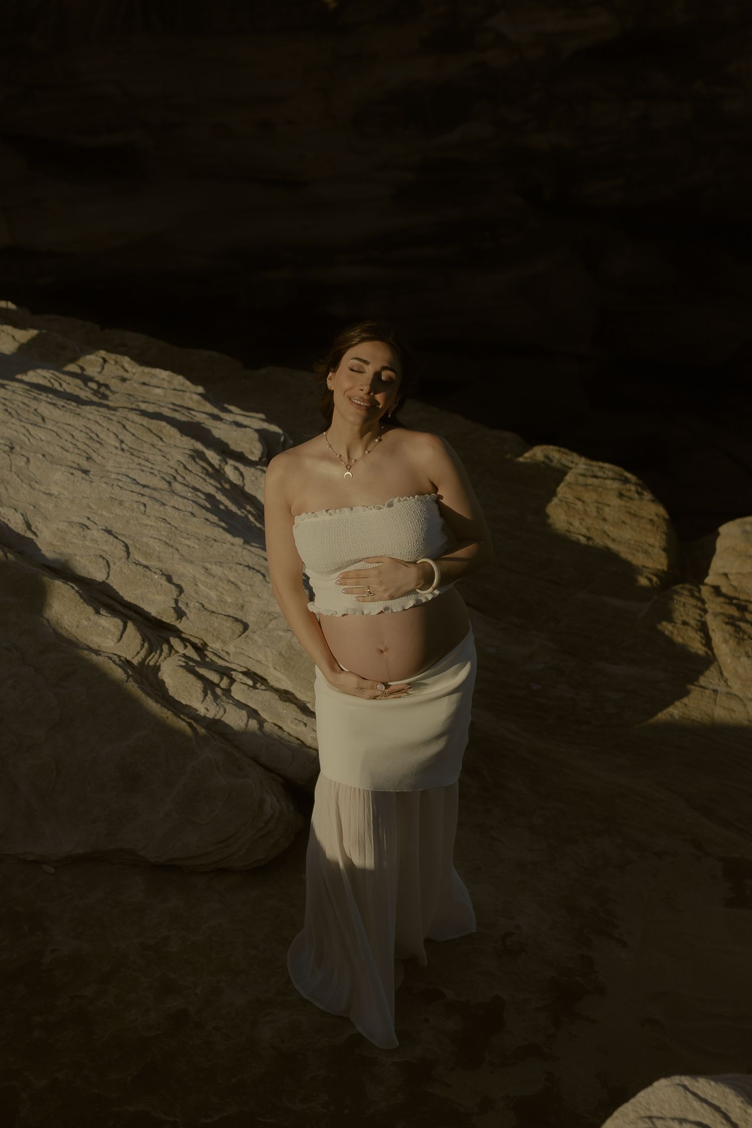 Pregnant woman in white two-piece maternity outfit poses gracefully among sunlit rocky formations.