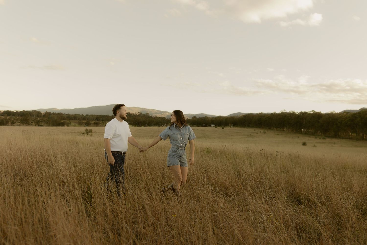 Couple holding hands strolling through tall golden grass field with mountain backdrop at sunset.
