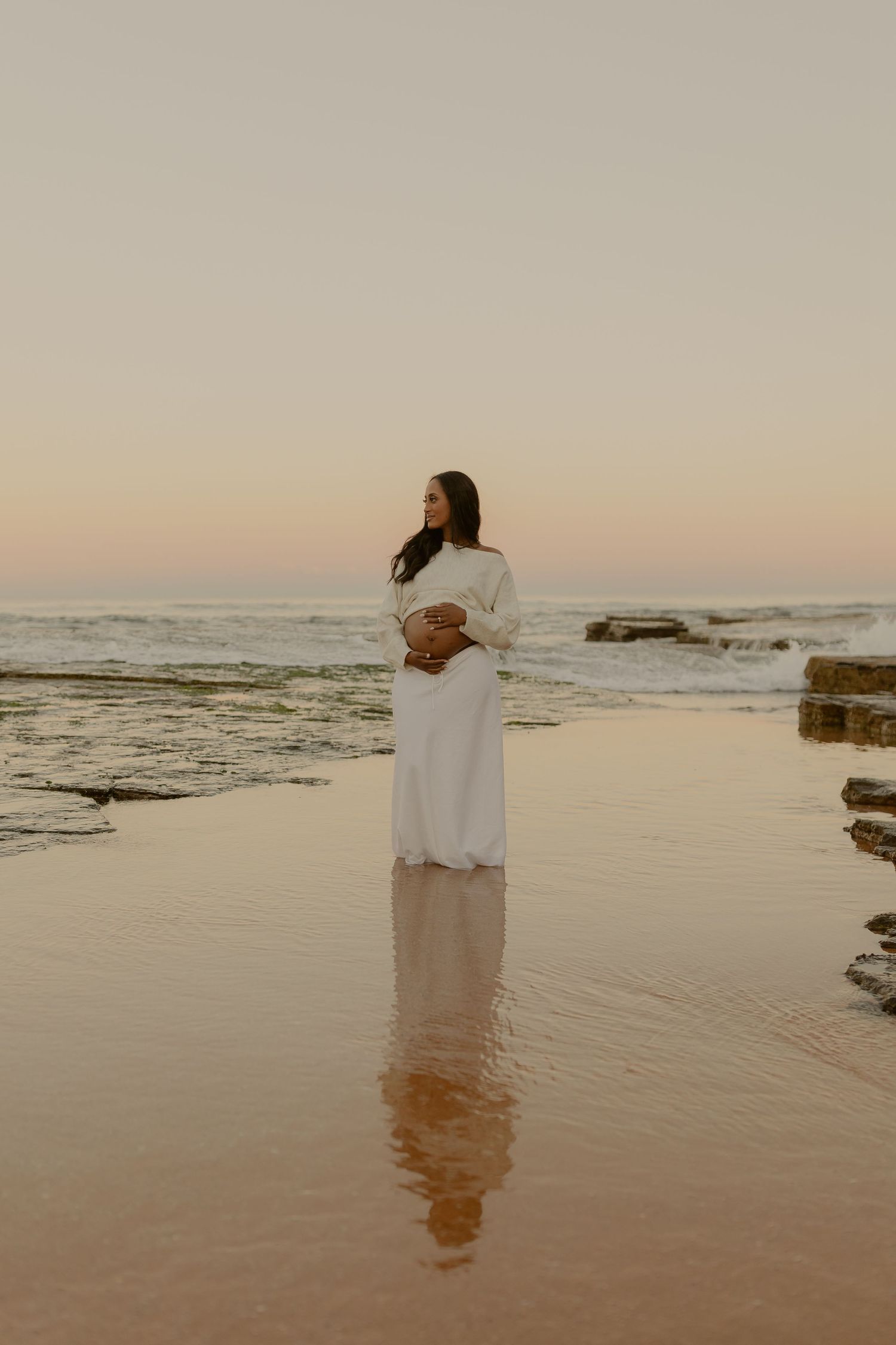 Pregnant woman in white dress standing on a reflective beach at sunset, cradling her baby bump near ocean waves.