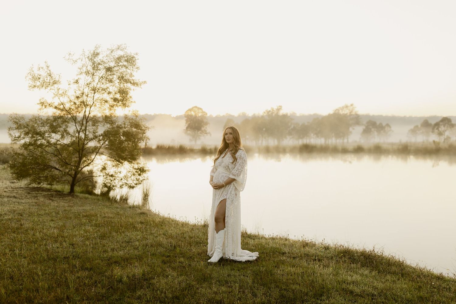 Pregnant woman in white gown standing by misty lake at golden hour sunrise in a serene outdoor maternity photo.