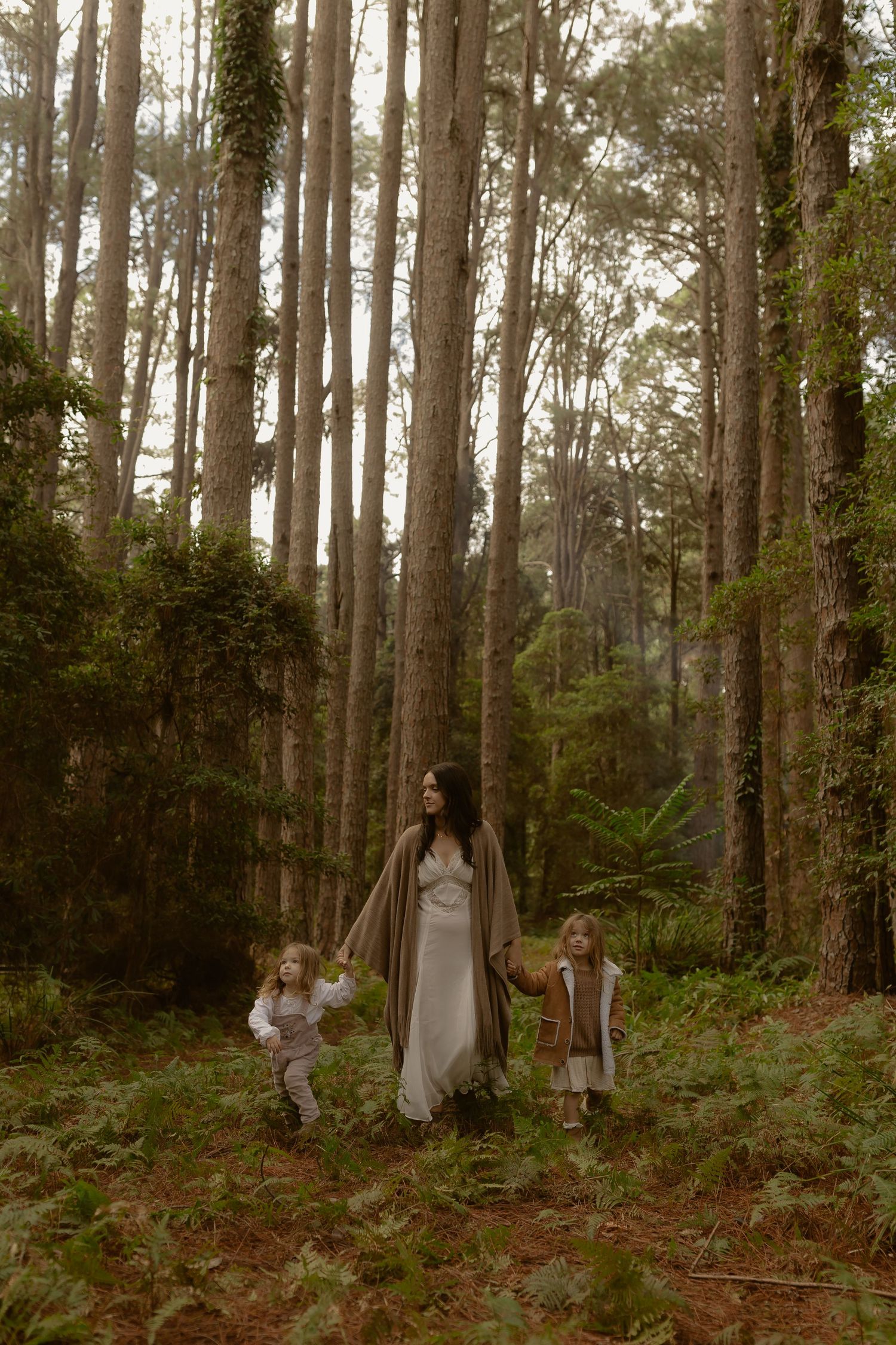 Mother in white dress walks with two young children through a lush forest setting.