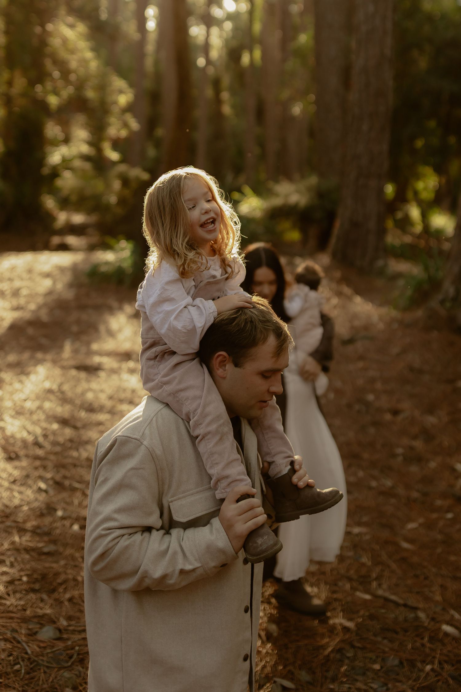 Father carries laughing daughter on shoulders in sunlit forest while mother holds baby.