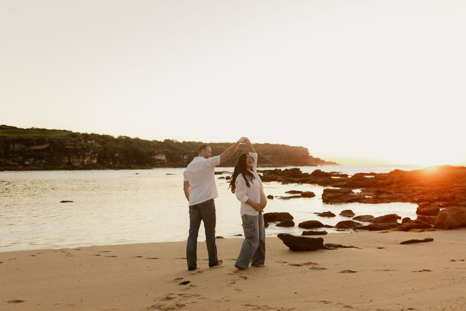 Couple standing together on sandy beach at sunset during golden hour maternity photoshoot.