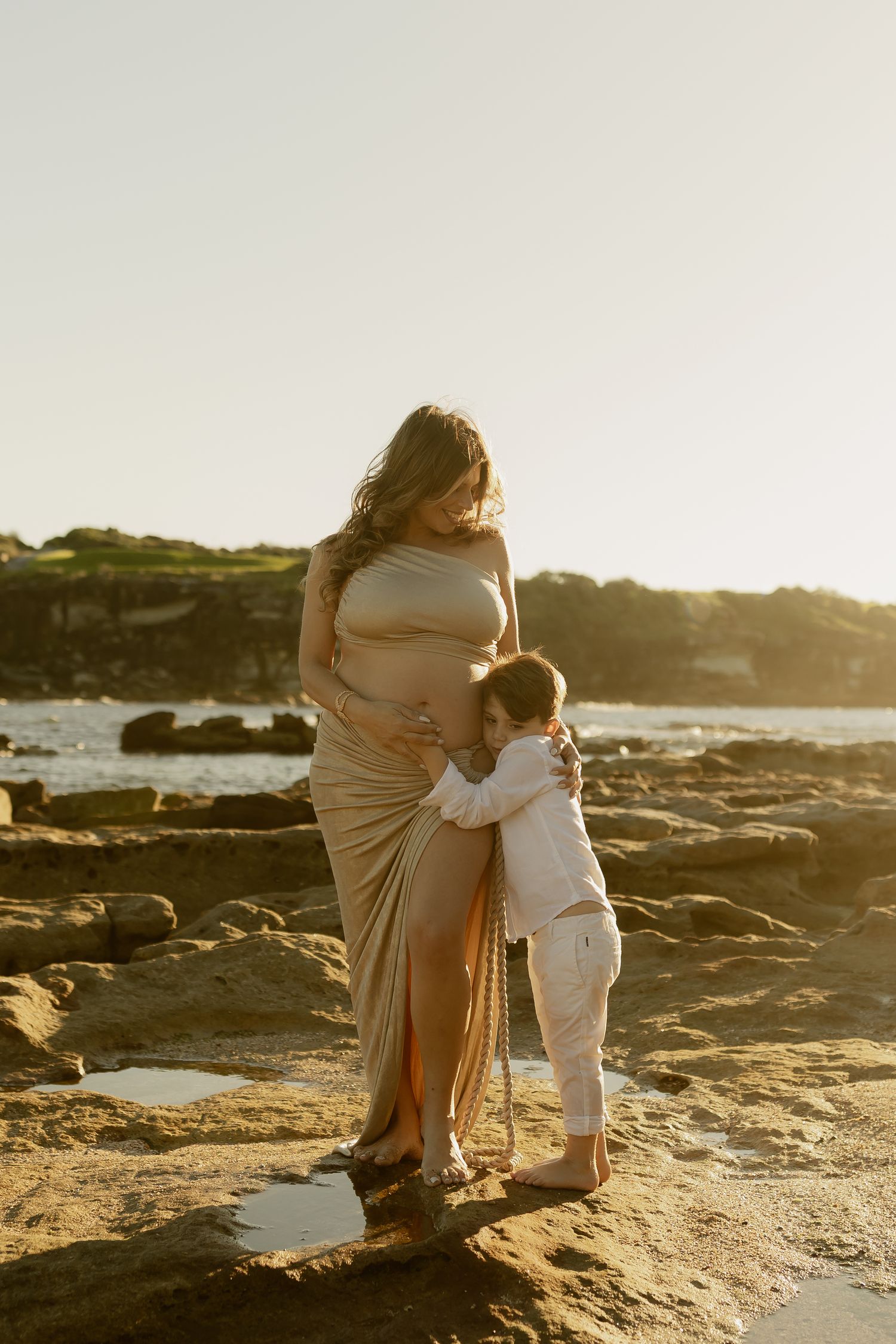 Pregnant woman in beige wrap dress hugged by young child on rocky beach at golden hour.