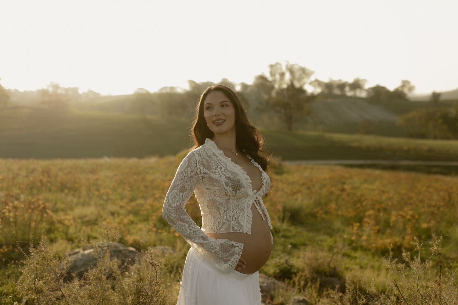 Elegant maternity portrait featuring woman in lace gown against sunset landscape.