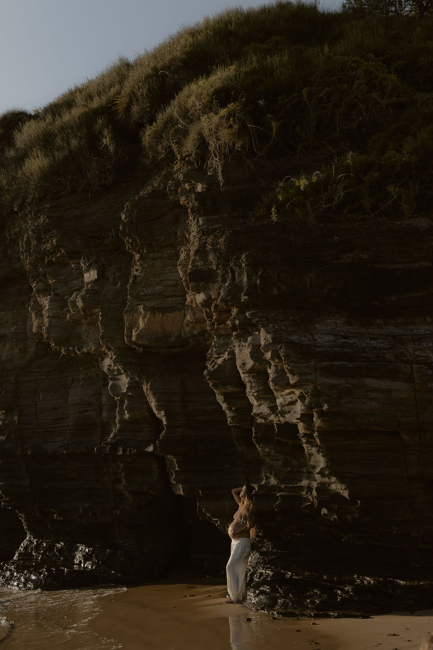 Silhouette of person in white standing near rugged coastal cliffs at sunset.