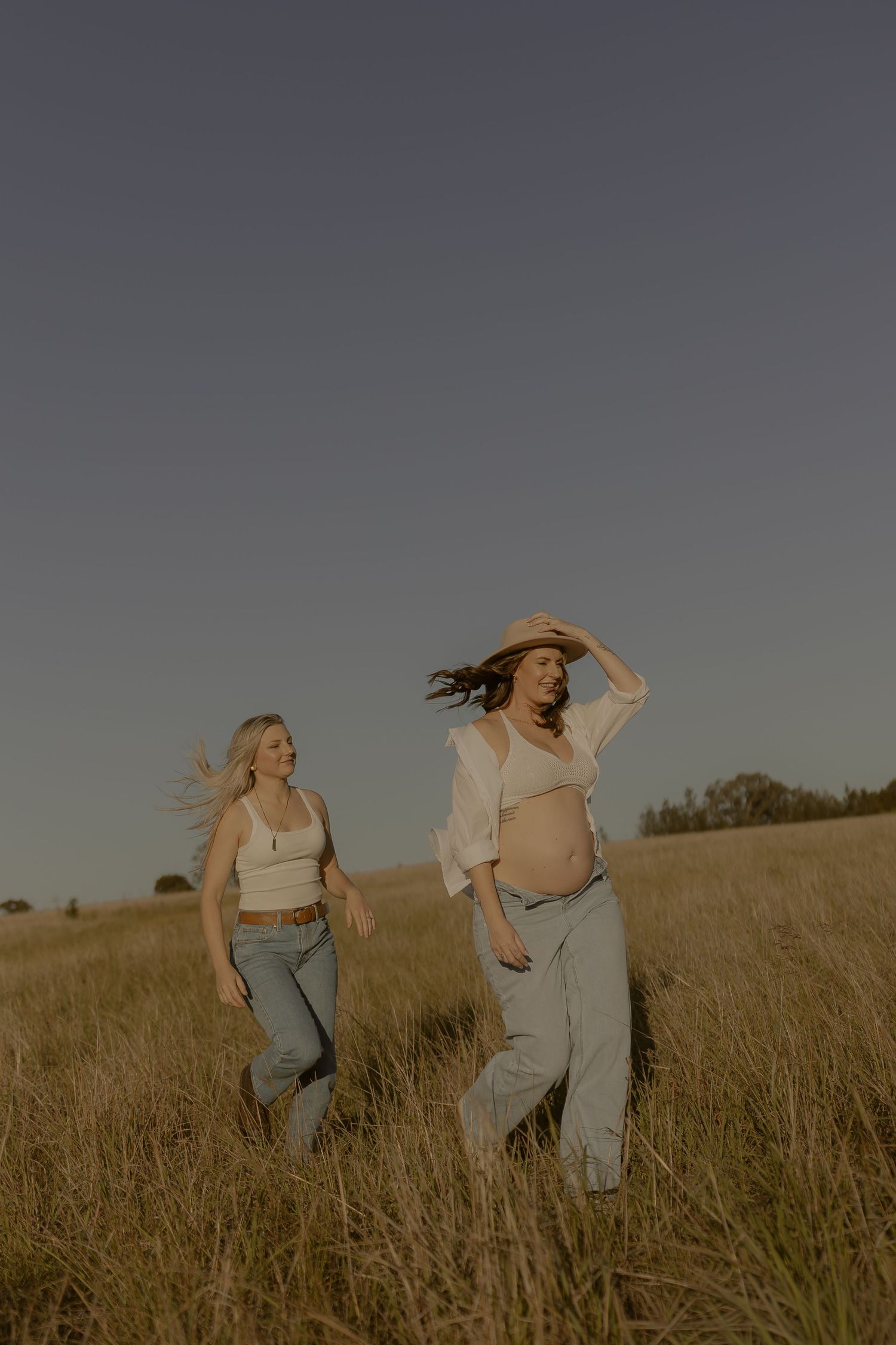 Two women in matching outfits run playfully through a golden field at sunset, their hair flowing in the wind.