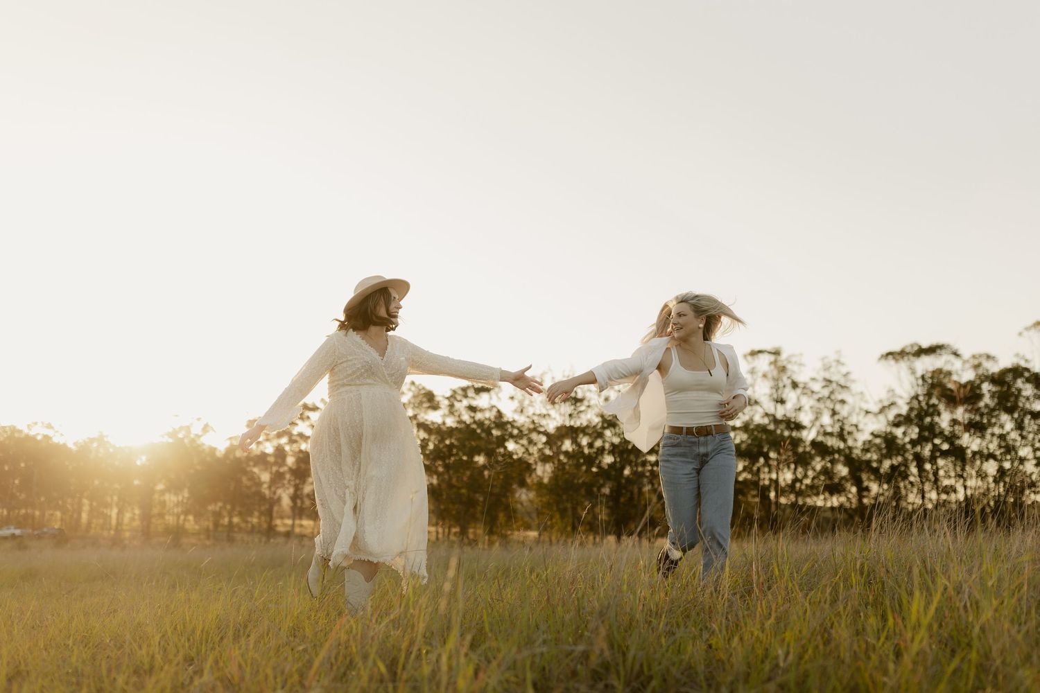 A couple walks hand in hand through a sunlit field at golden hour with their dog beside them.