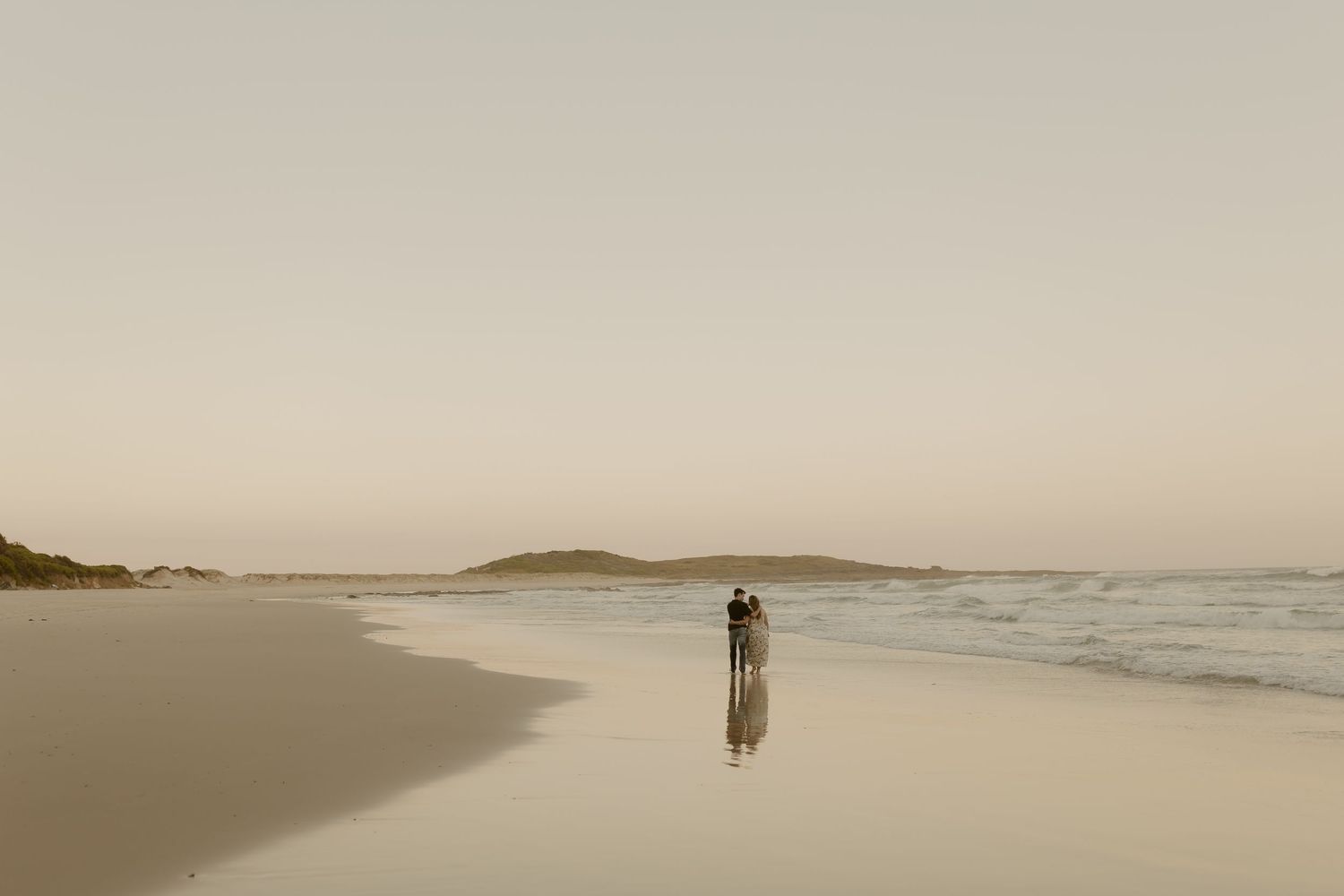 A solitary figure stands on a serene beach at dusk with gentle waves and distant islands on the horizon.