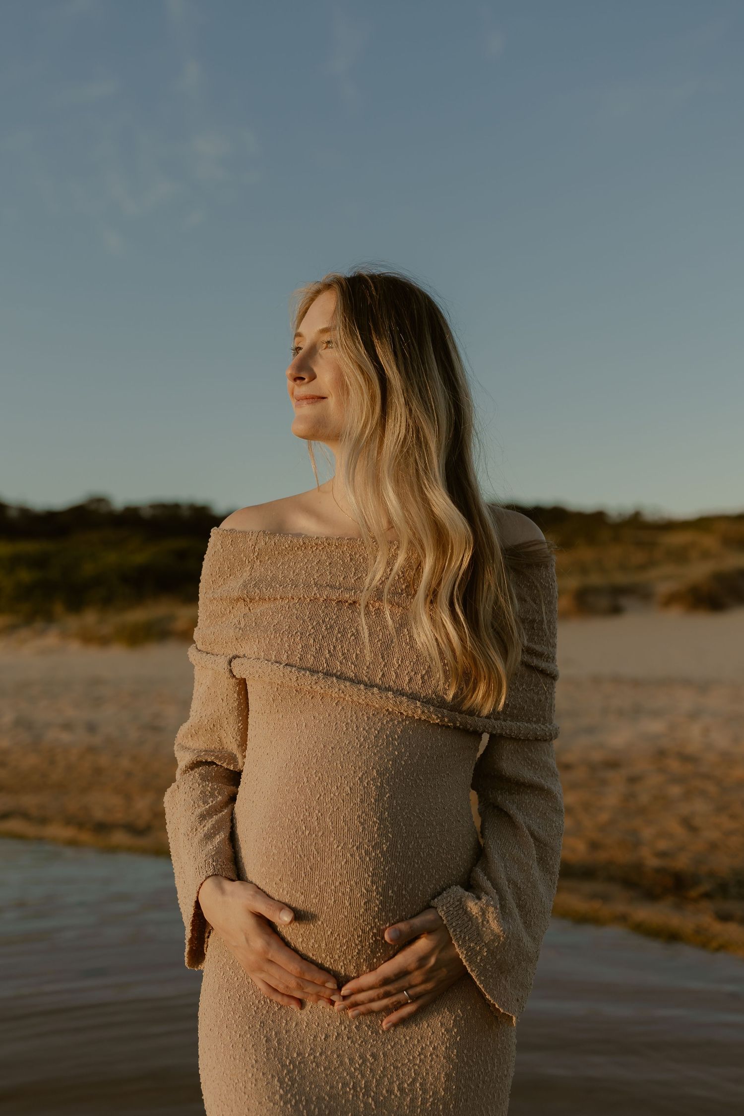An expectant mother in a beige sweater dress poses outdoors during golden hour with a peaceful expression.