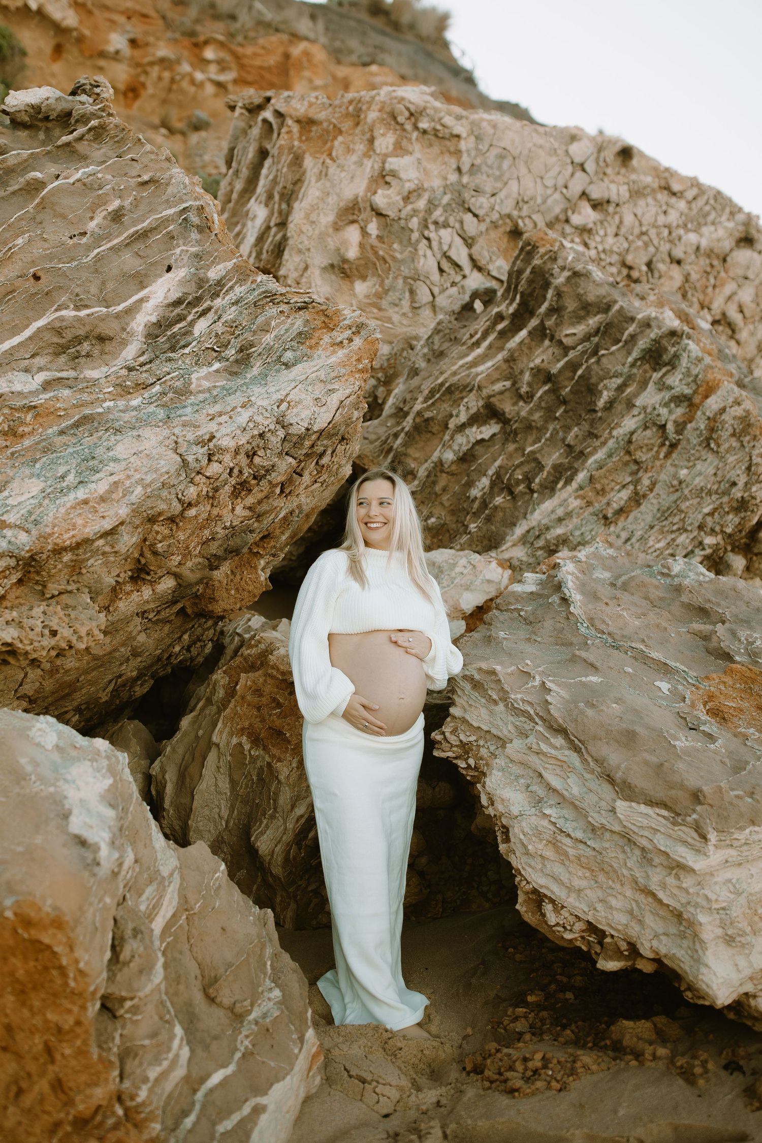 A pregnant person wearing a white dress poses between large rock formations during a maternity photoshoot.