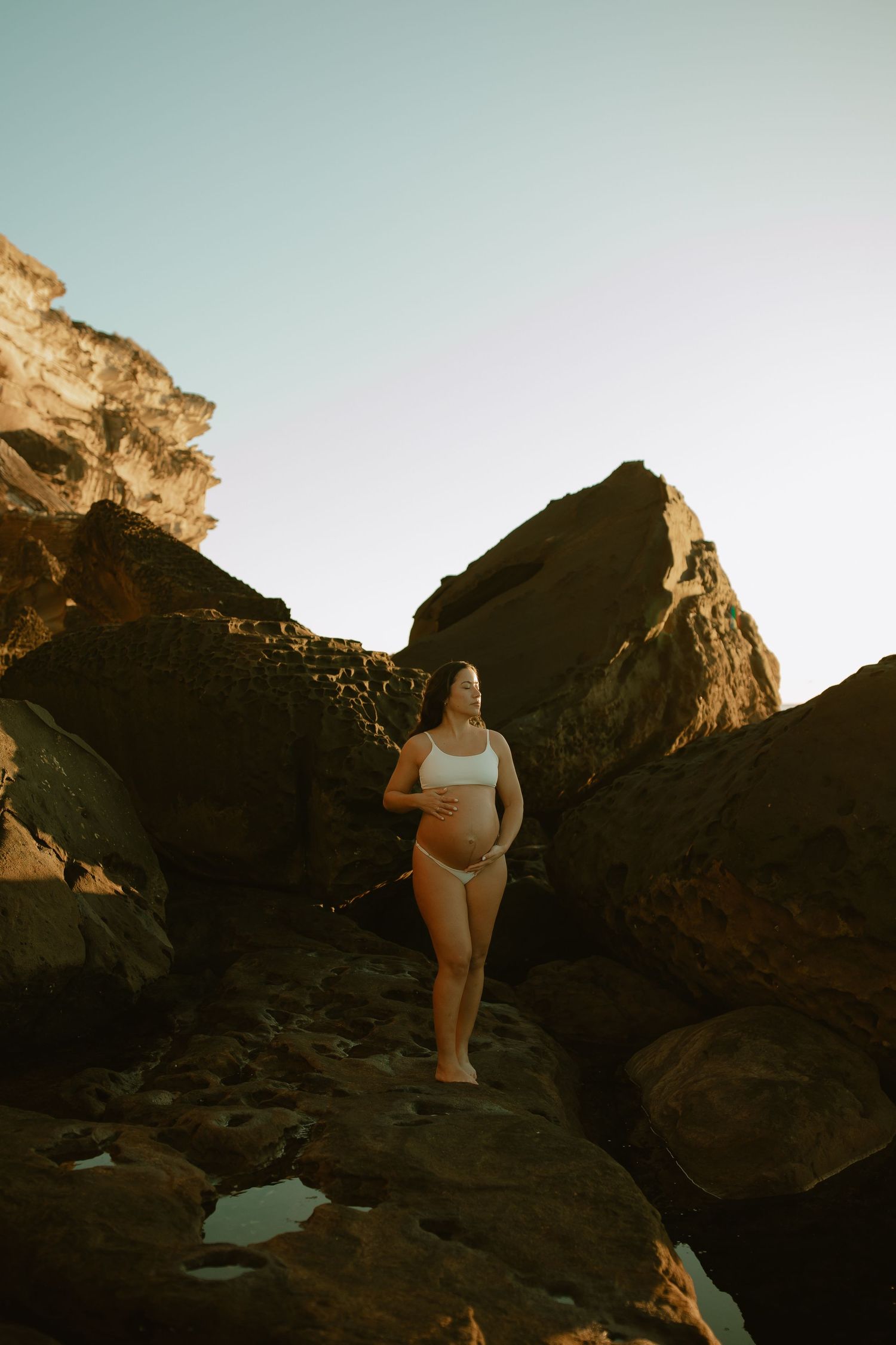 A person in white swimwear poses on rocky cliffs at sunset overlooking a scenic natural landscape.