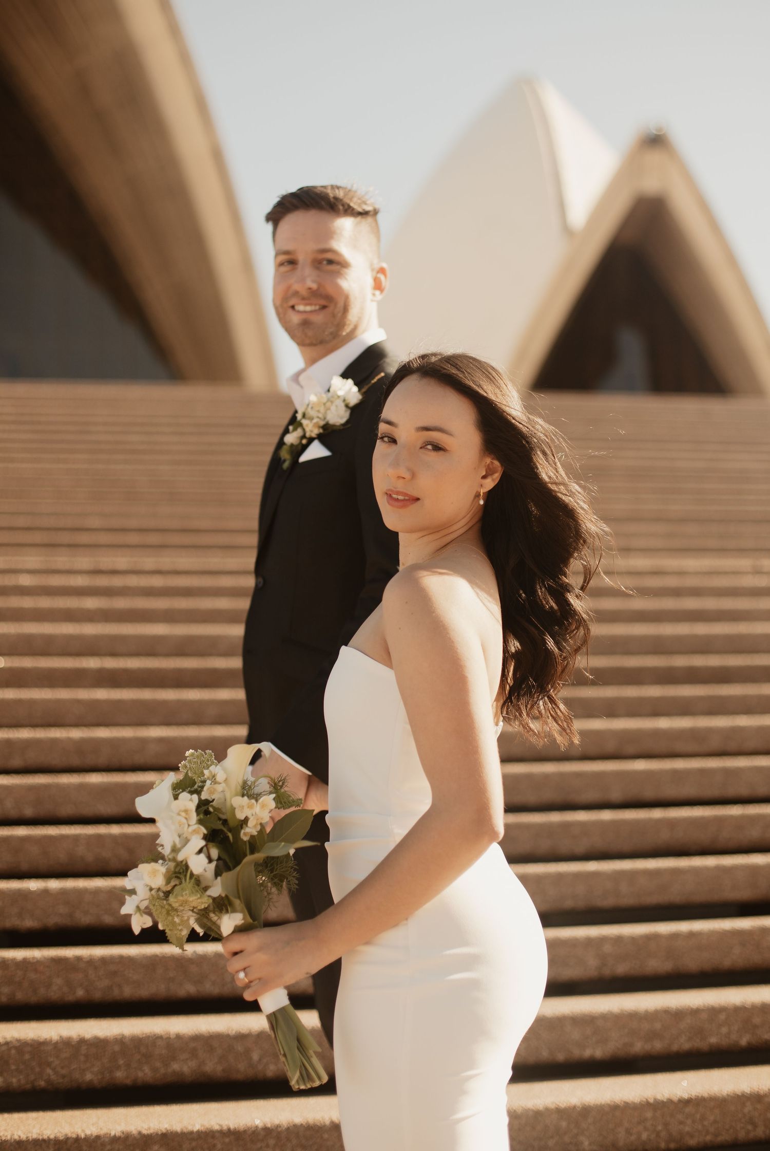 A couple poses on the steps of the Sydney Opera House during their wedding photoshoot at sunset.