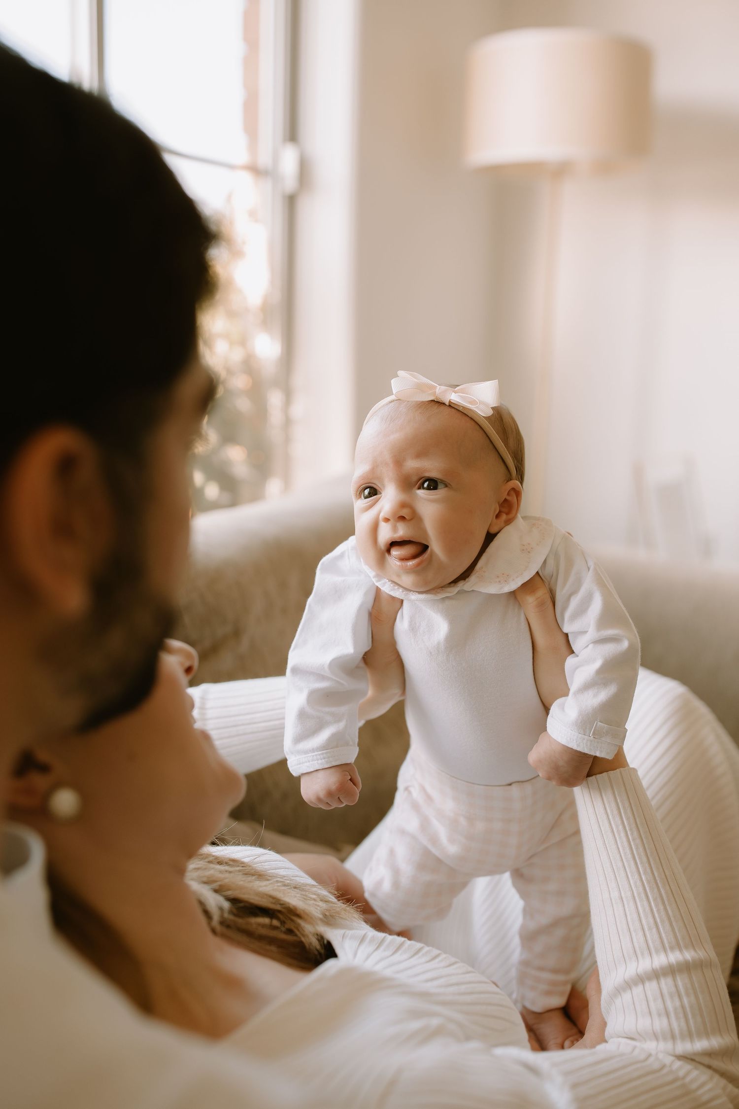 Smiling baby in white outfit with bow headband stands on soft bedding while caregiver looks on in warm sunlit room.