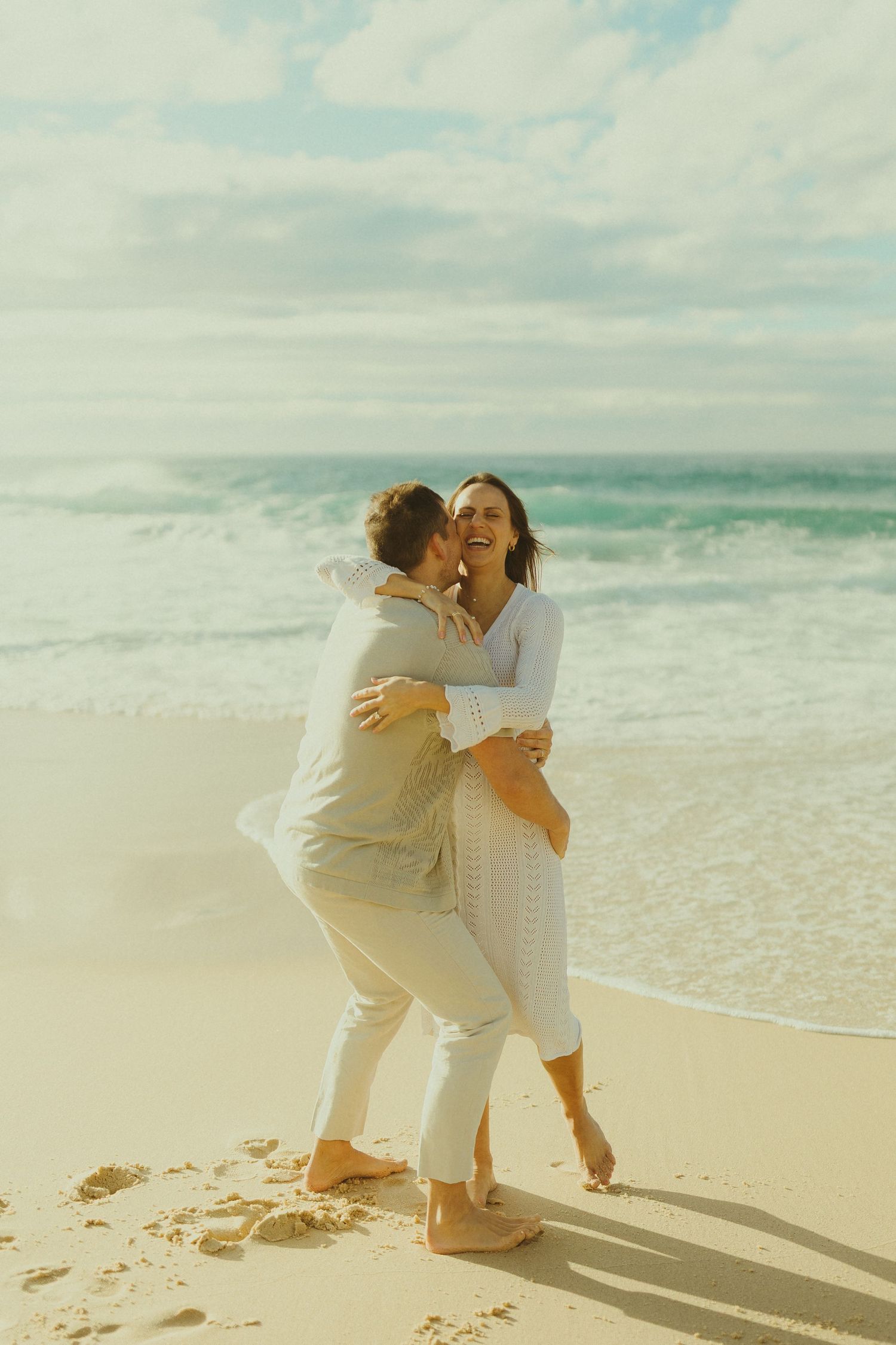 A couple embraces joyfully while wearing white outfits on a sun-drenched beach with ocean waves in the background.