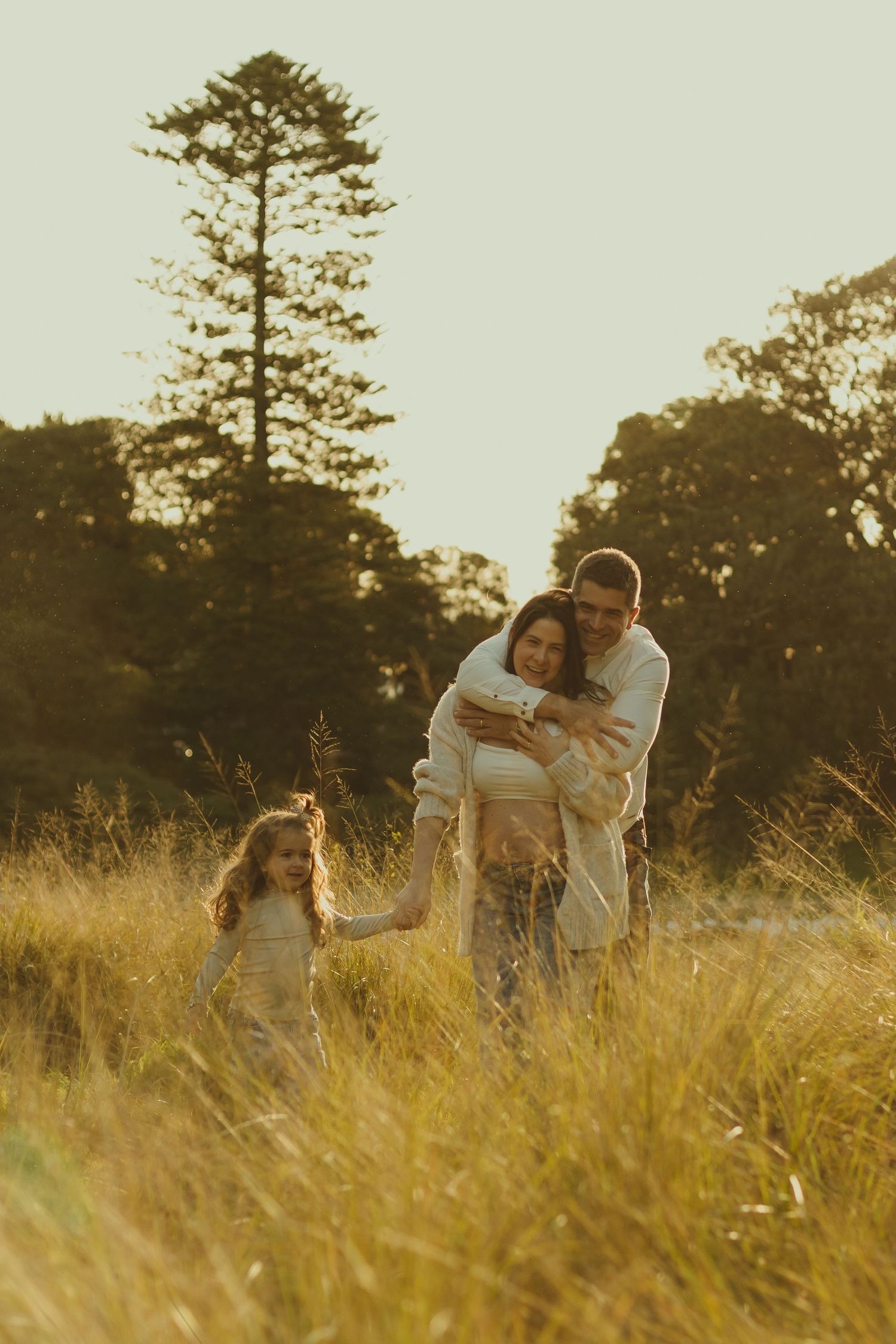 Family walking through tall golden grass at sunset with towering pine tree in background.
