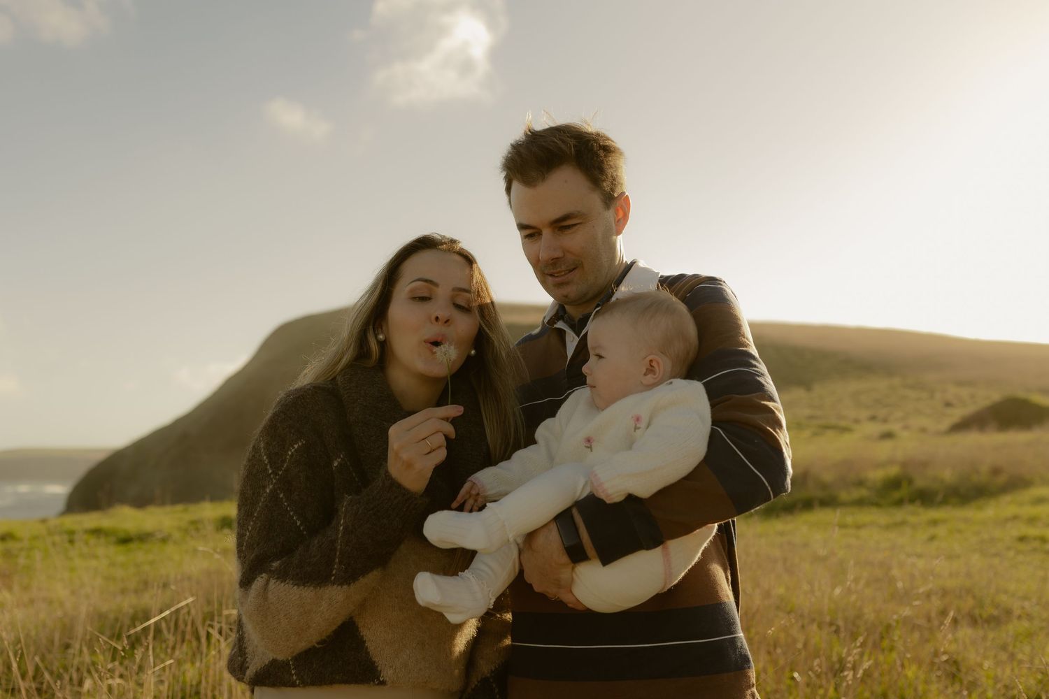 Young couple holding baby outdoors on green hillside at golden hour with coastal cliffs in background.