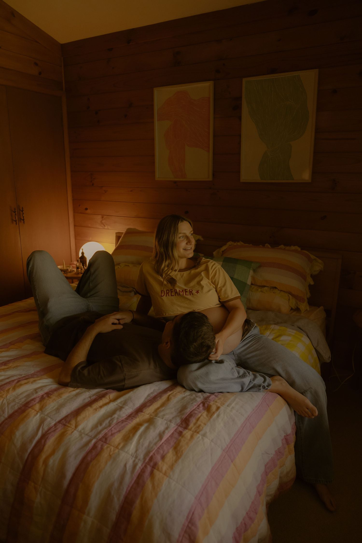 A pregnant woman smiles warmly while relaxing on a striped bed with her partner in a cozy wooden room.