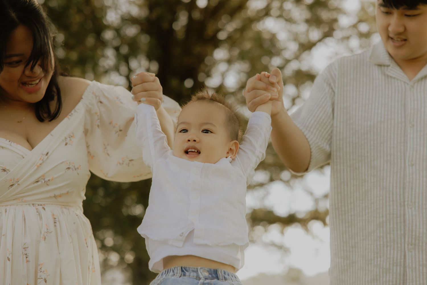 Happy baby held by hands between two parents outdoors in a bright family photo session.