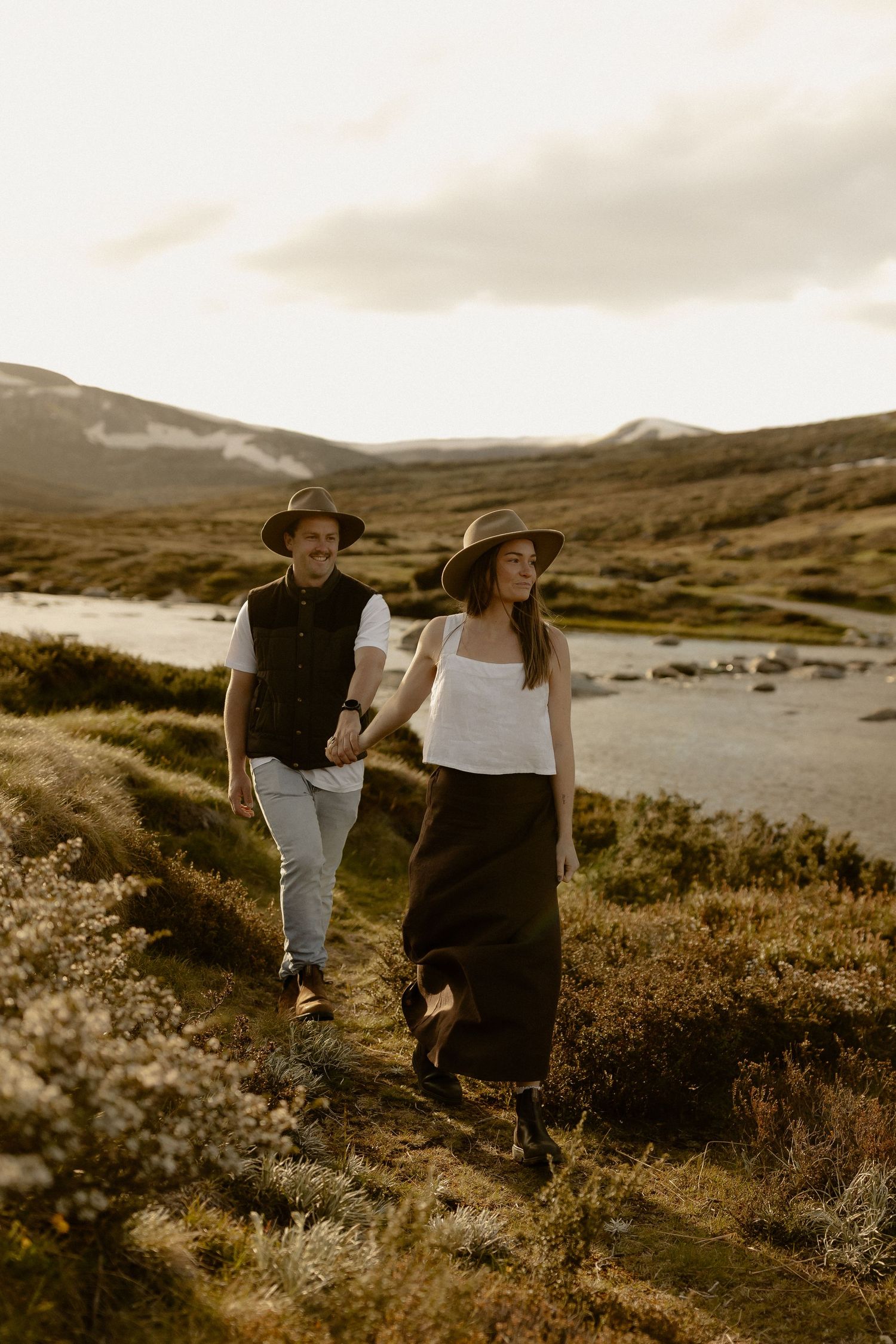 Two people in hats explore a rugged mountain terrain with wildflowers during golden hour.
