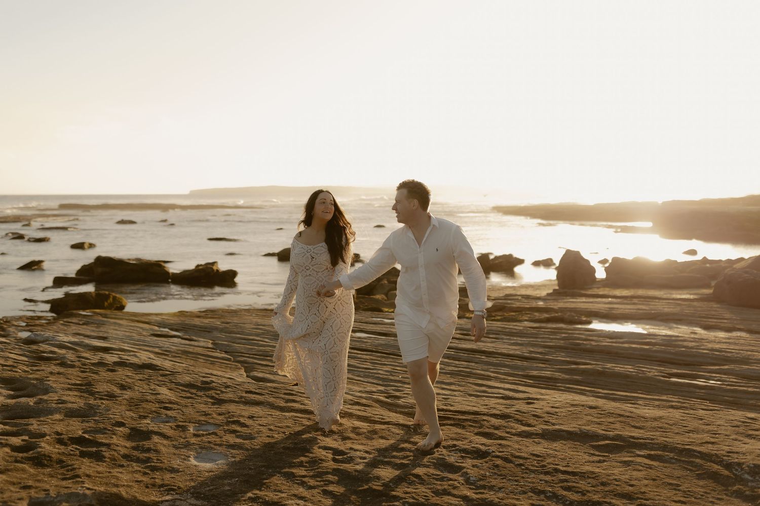 A couple in white outfits walks along scenic coastal rocks at sunset, creating romantic silhouettes against the golden sky.