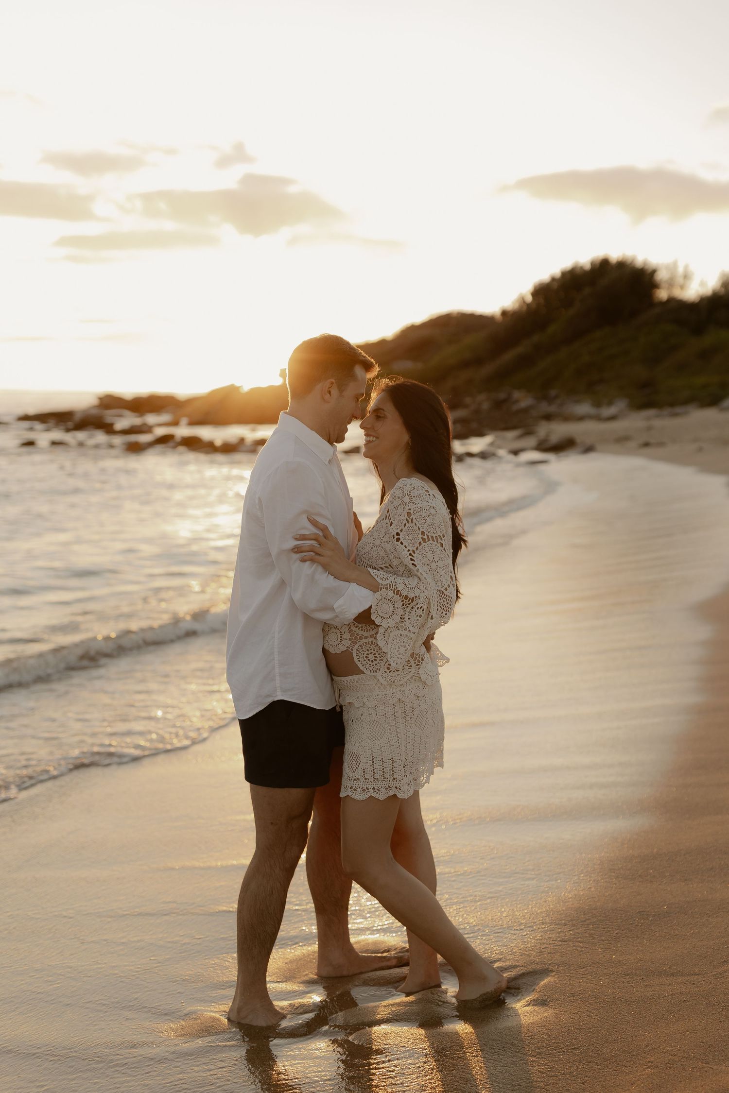 A romantic couple embraces on a sandy beach at sunset while ocean waves roll in along the shoreline.