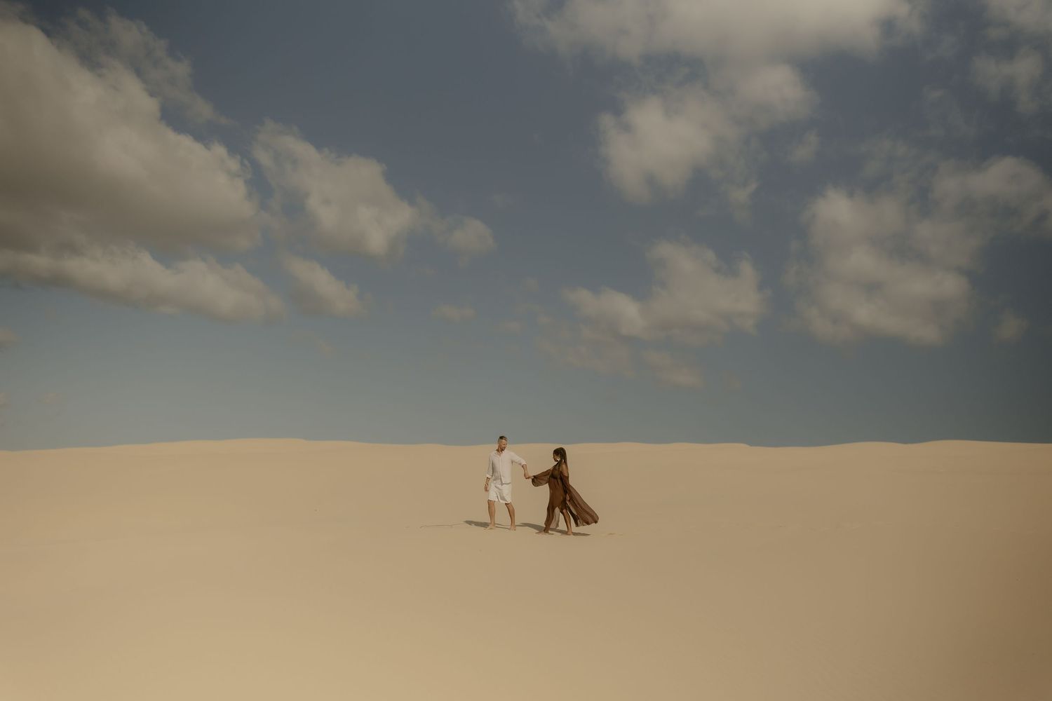 Two silhouetted figures walking together across vast sand dunes under a cloudy desert sky.