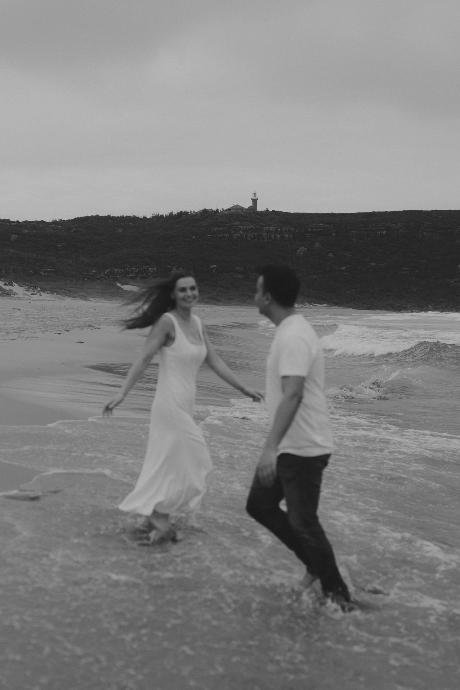 Two people in white and dark clothing run playfully along a beach shoreline in black and white photography.